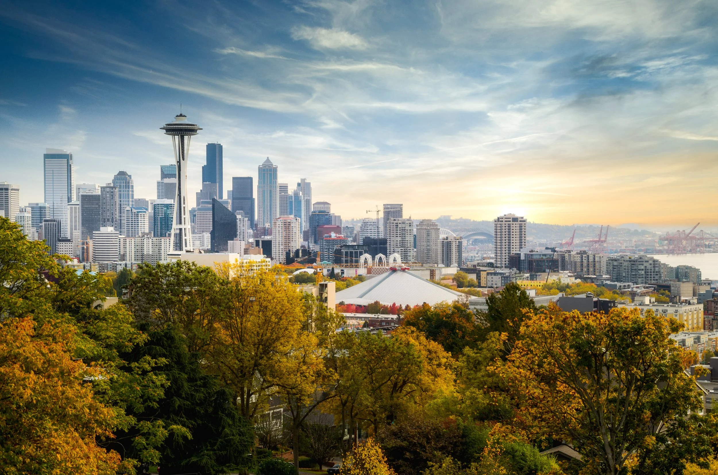 A city skyline with tall buildings, including the Space Needle, under a partly cloudy sky with sunlight, with colorful fall trees in the foreground.
