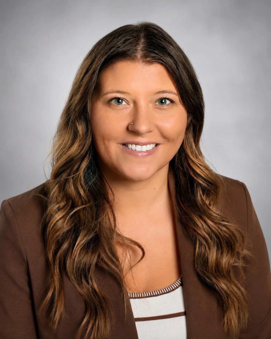 Headshot of a smiling woman with long, wavy brown hair, wearing a brown blazer over a white top with brown stripes, against a plain gray background.