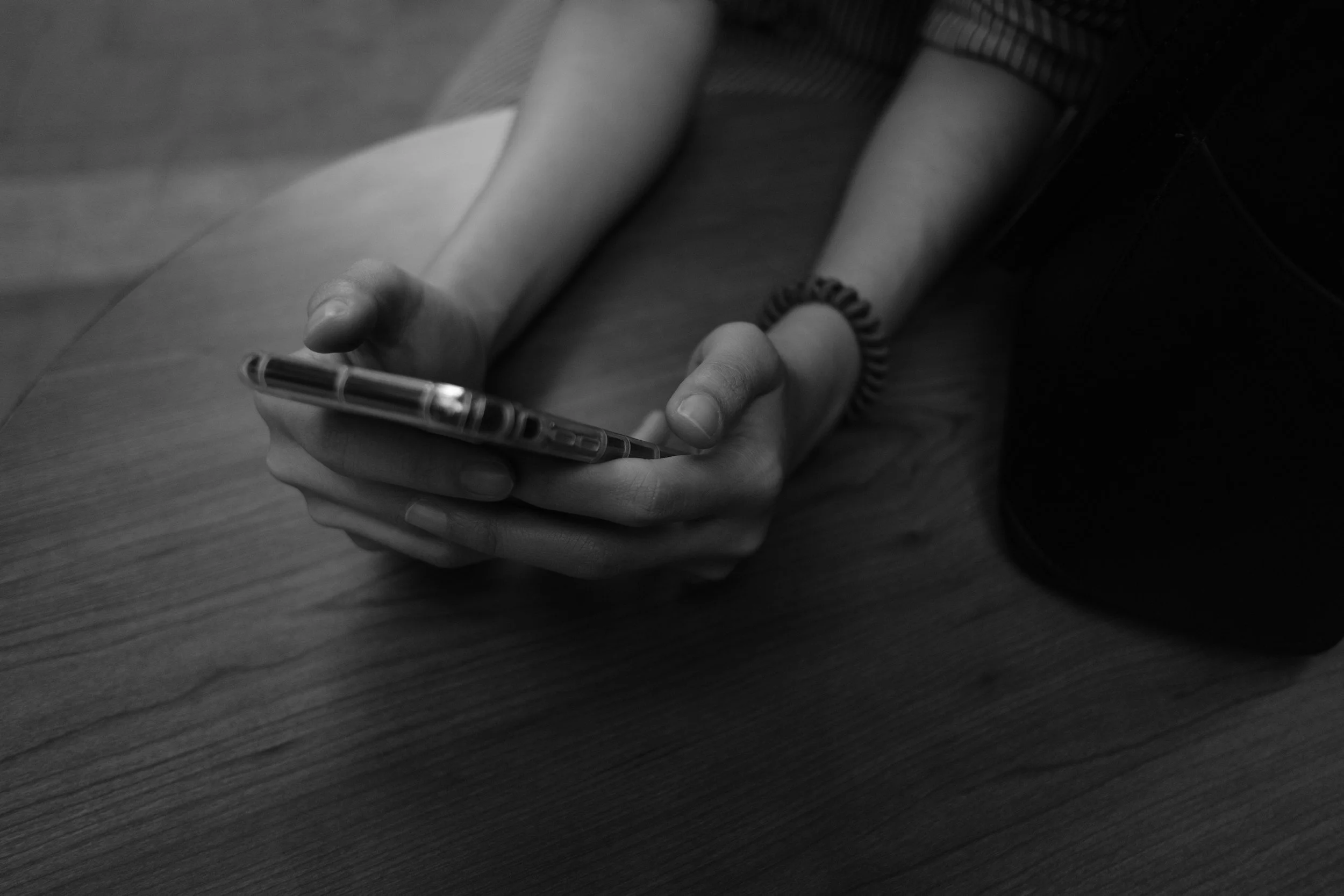 A person holding a smartphone with both hands on a wooden table, wearing a wristband and a bracelet.