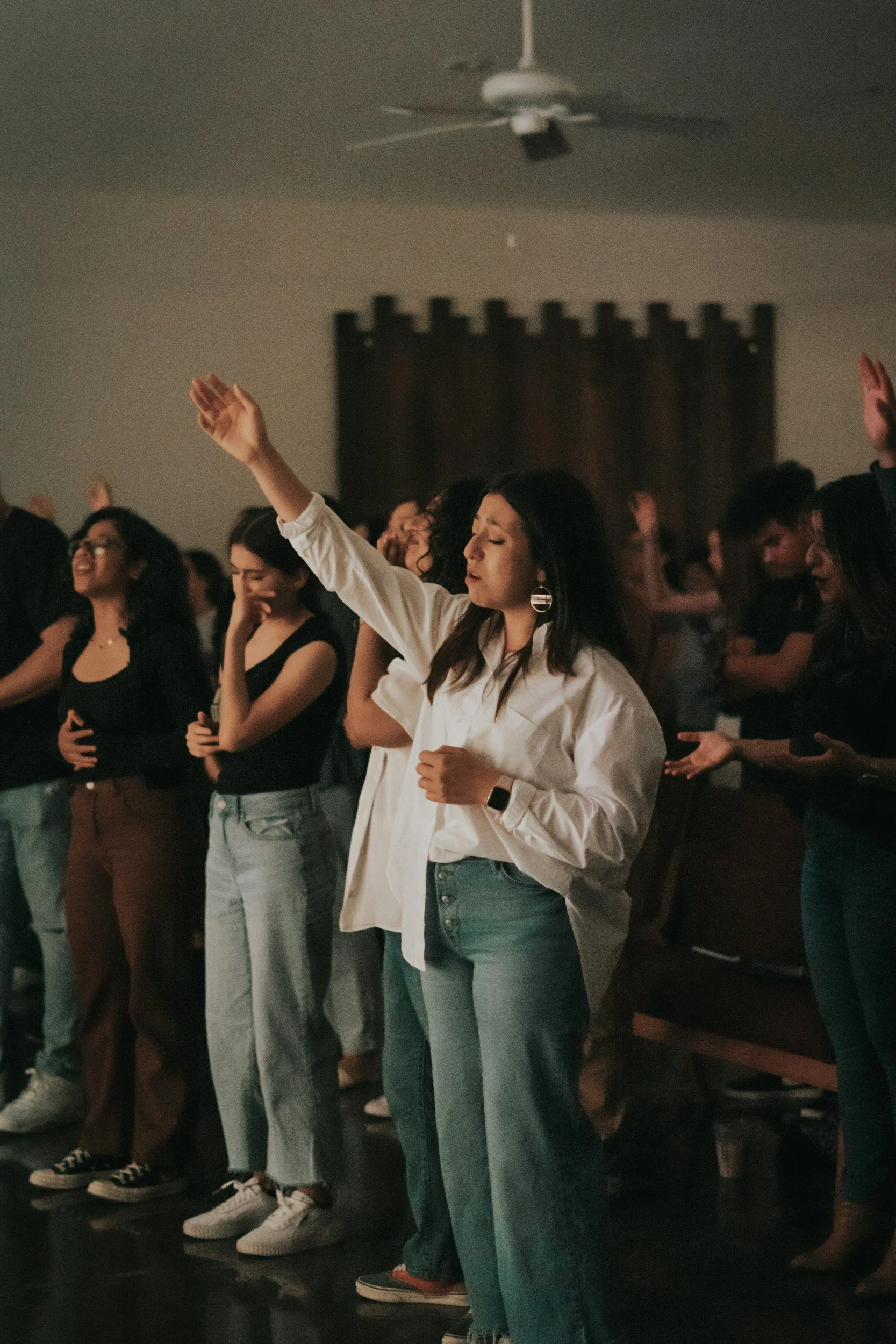 A group of people, mostly young women, standing with eyes closed, some with hands raised, in a dimly lit room during a worship or prayer session.