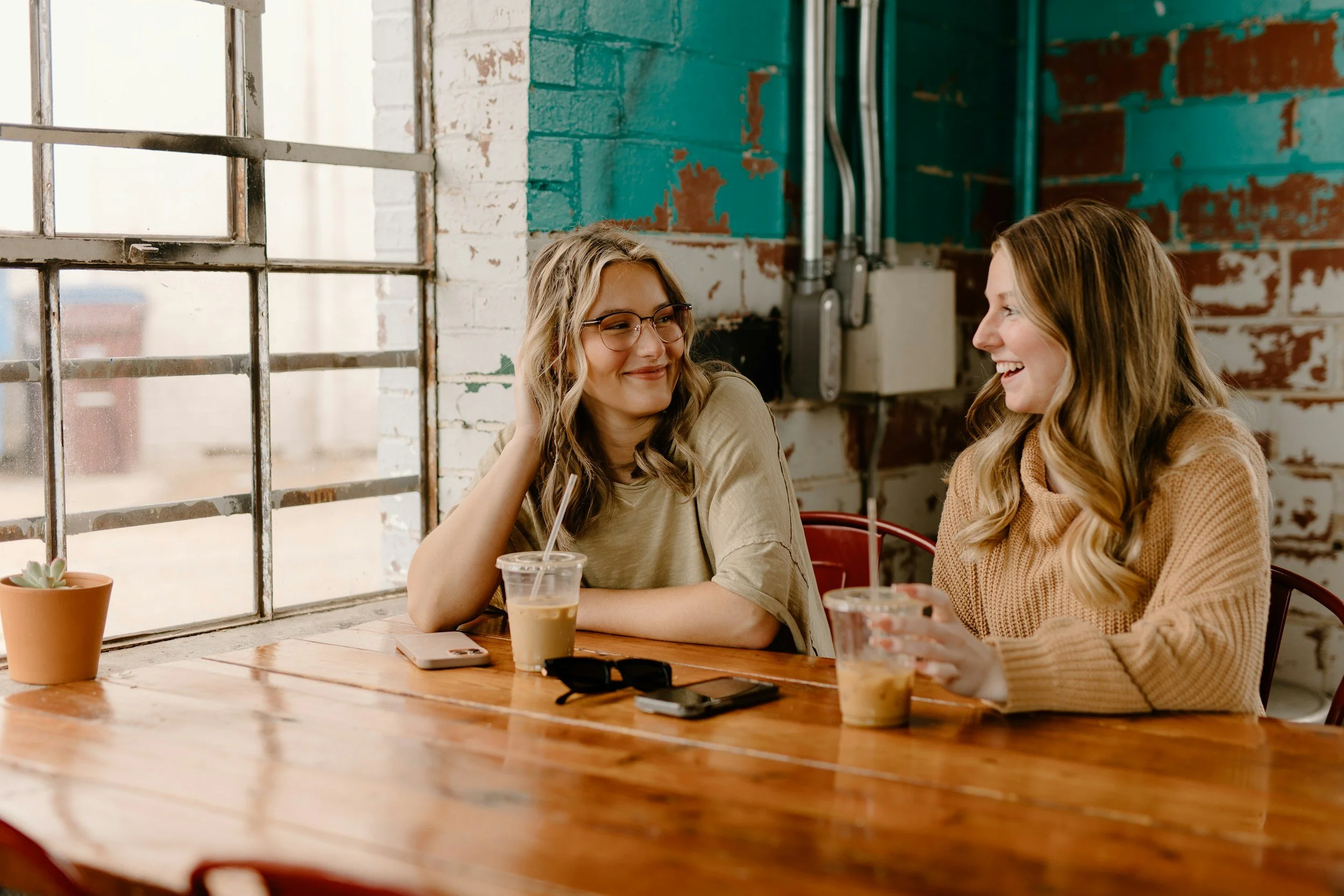 Two women sitting at a wooden table in a cafe, smiling and talking, with drinks in front of them and a brick wall with pipes behind.