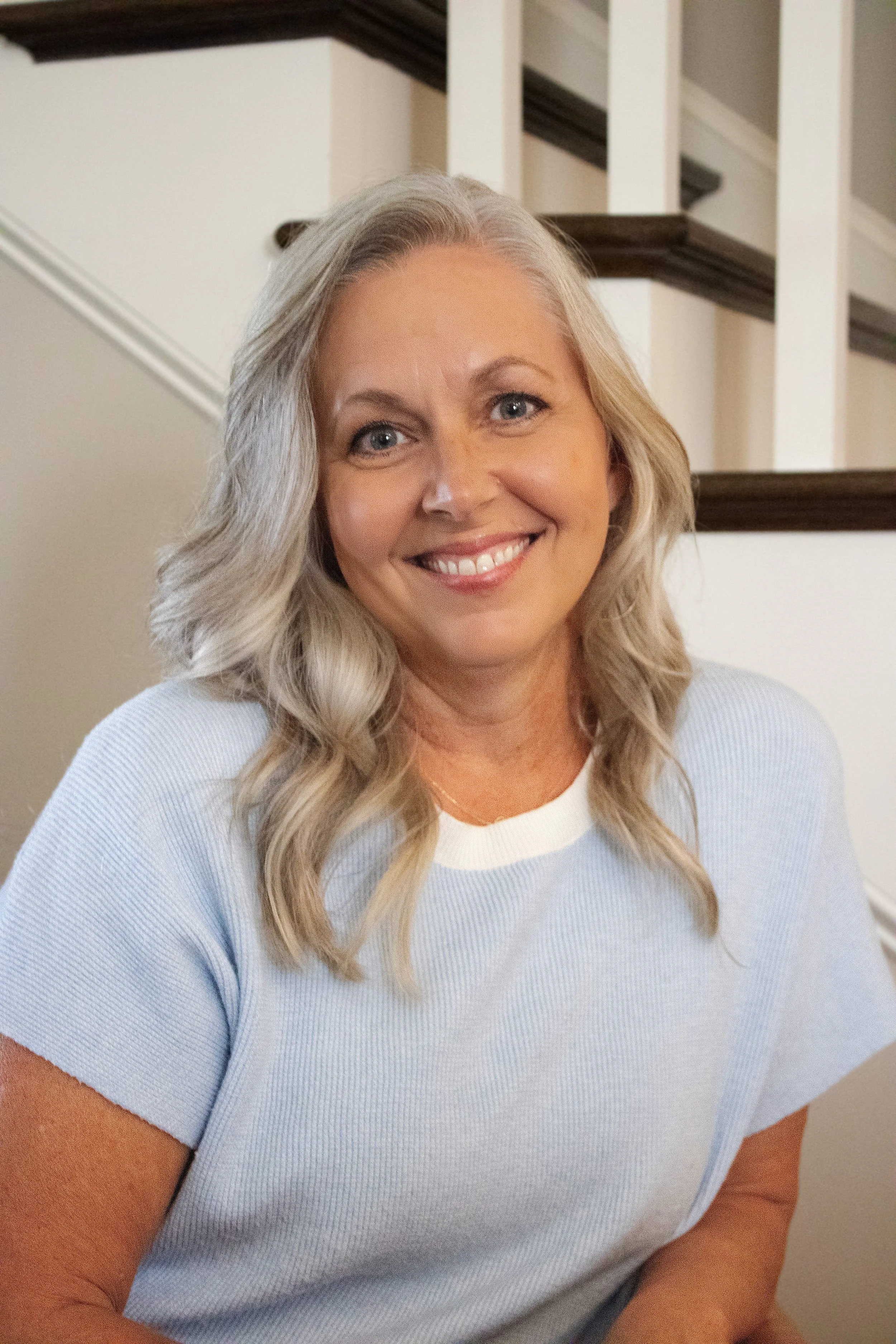 A woman with blonde hair smiling and wearing a light blue top, sitting indoors near a staircase.
