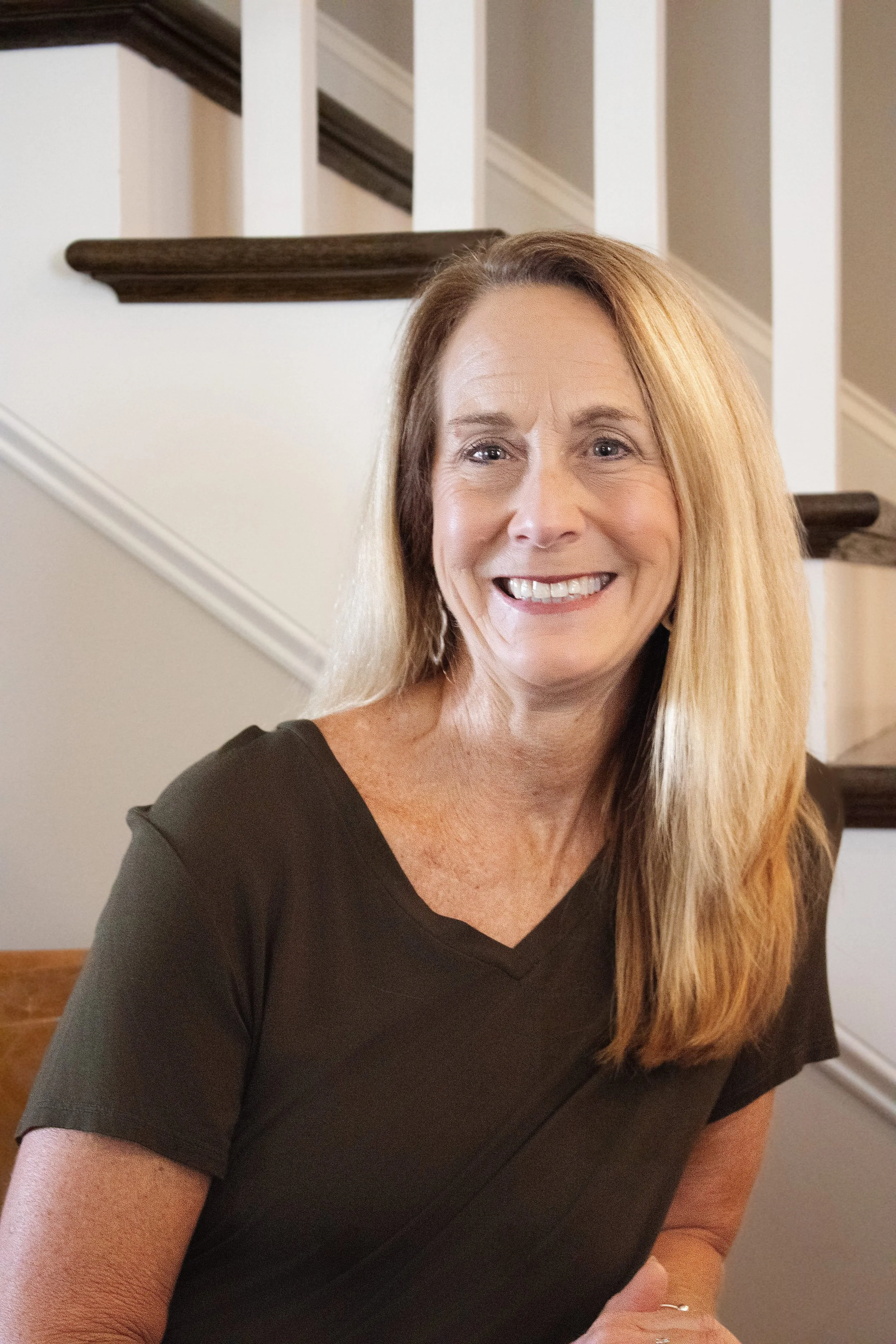 A woman with long blond hair smiling at the camera, sitting indoors near a staircase with white spindles and dark wood handrails.
