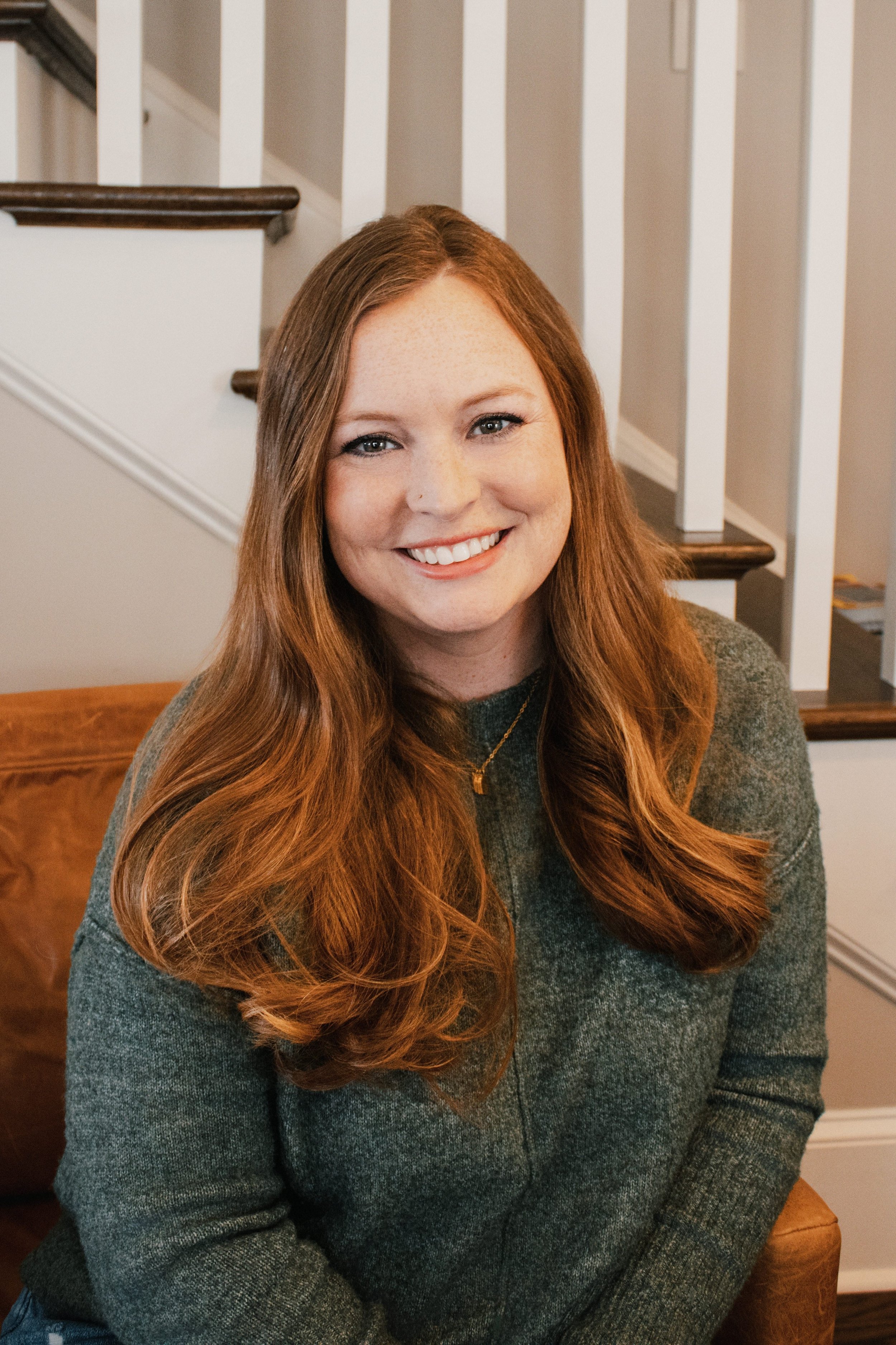 A woman with long red hair smiling outdoors, wearing a blue blazer and hoop earrings, with a green leafy background.