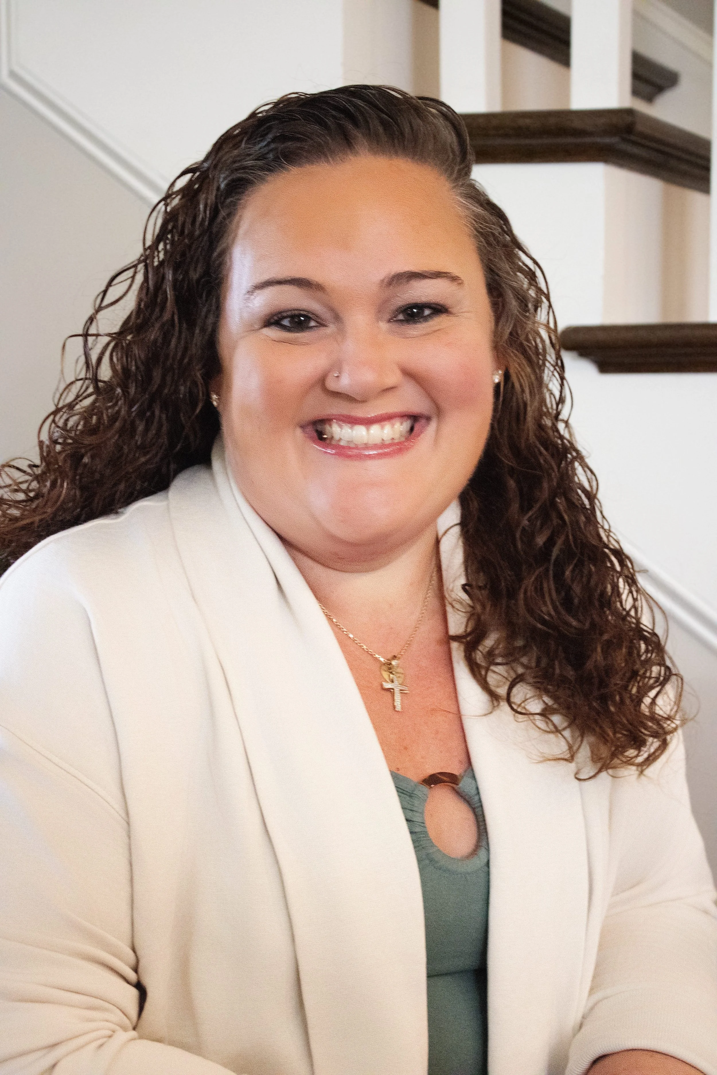 A woman with curly brown hair smiling, wearing a white blazer, green top, and a gold cross necklace, sitting indoors near a staircase.