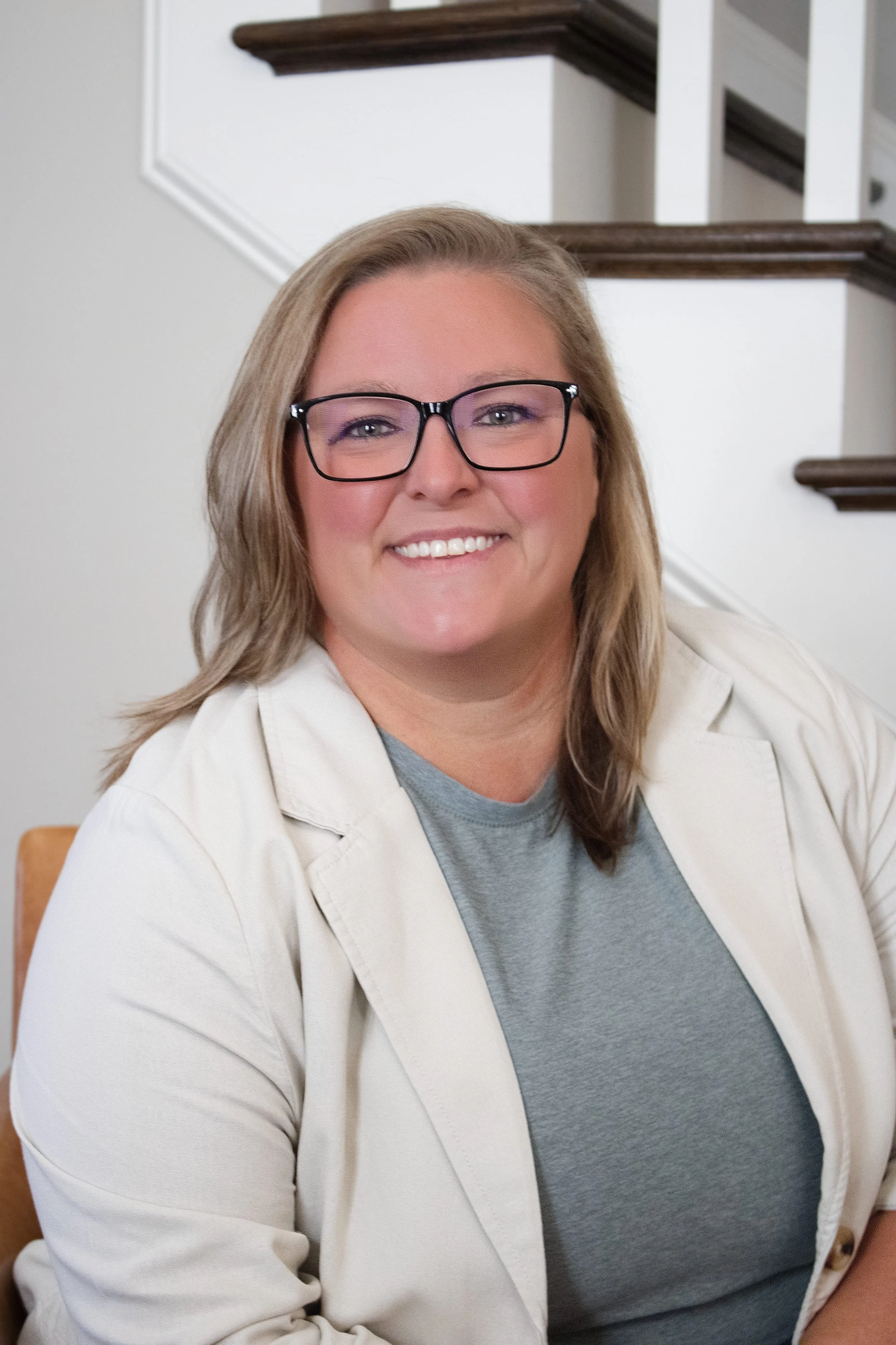 A woman with blond hair, glasses, and a gray shirt, smiling at the camera, seated indoors against a staircase.