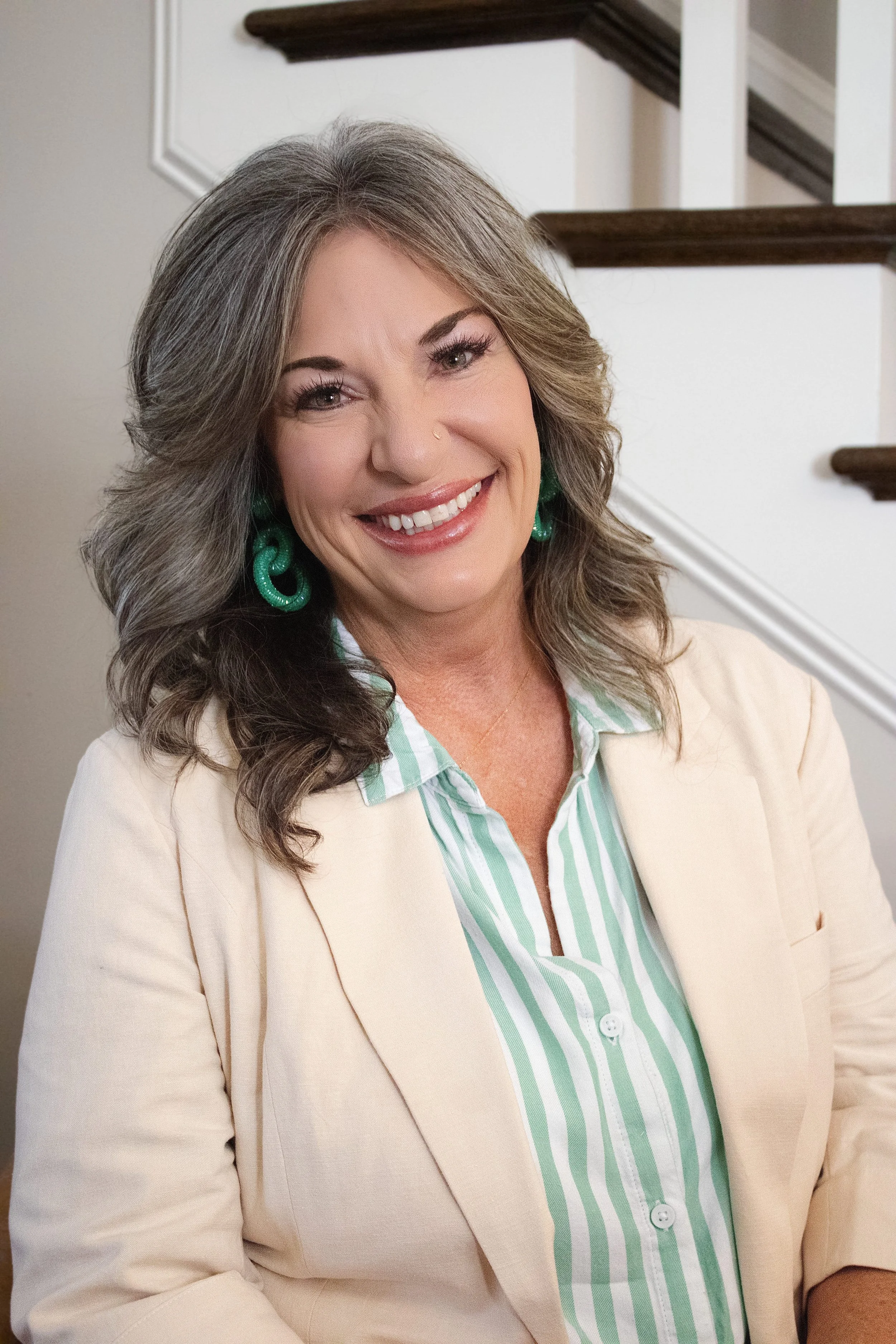 A smiling woman with gray hair, wearing a beige blazer, green earrings, and a green-and-white striped shirt, standing indoors near a staircase.