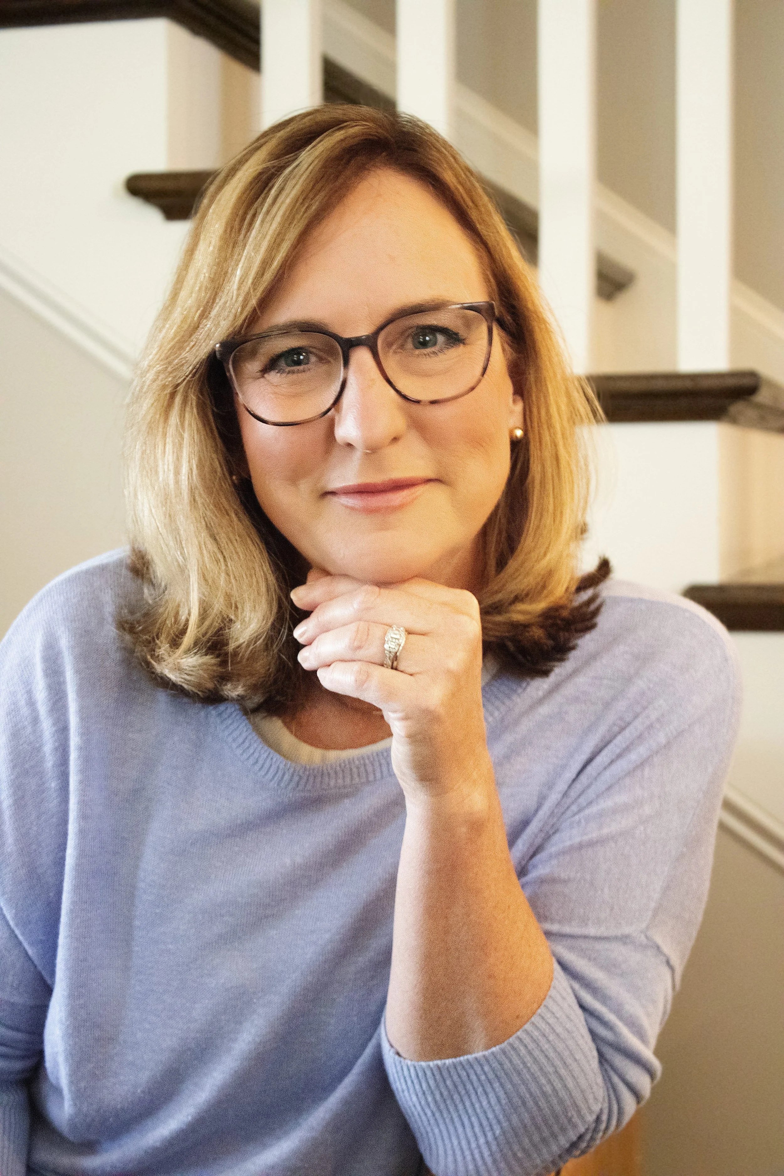 A woman with shoulder-length blonde hair, glasses, and a light gray sweater, sitting indoors with a staircase in the background, resting her chin on her hand and smiling at the camera.