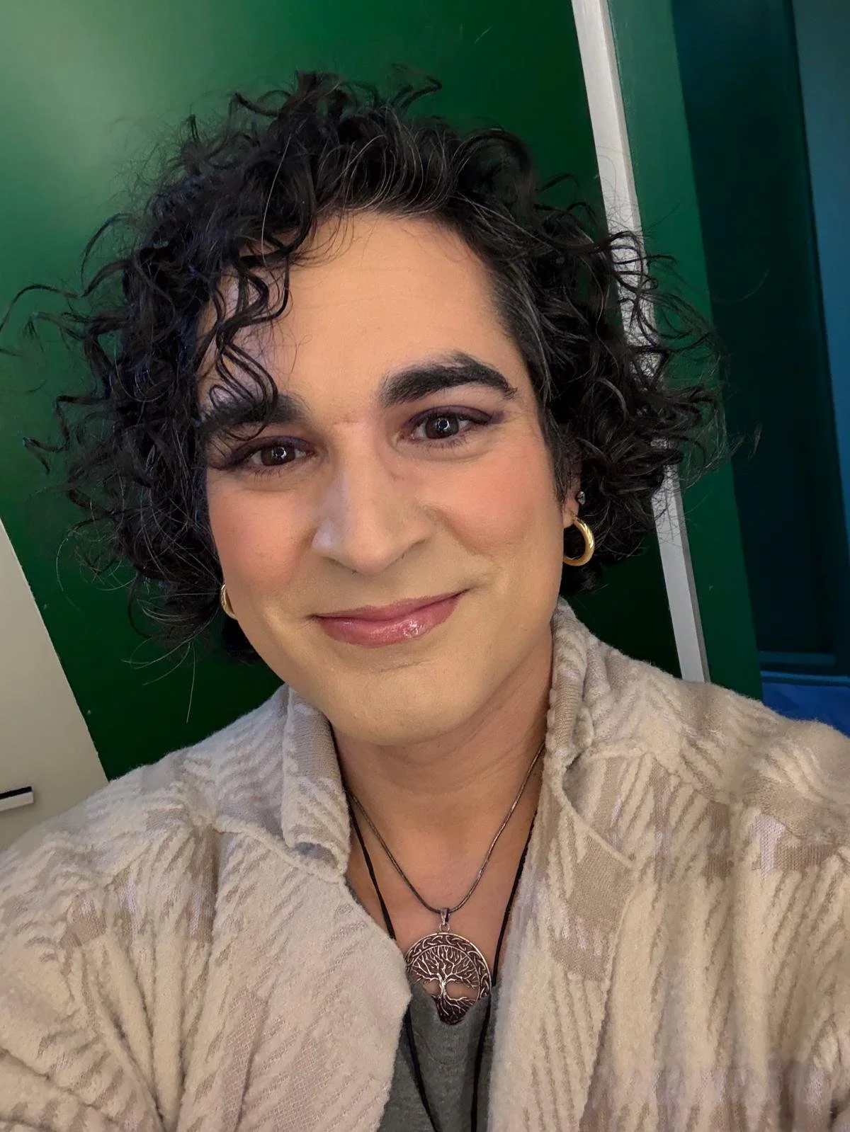 A woman with curly dark hair, wearing gold hoop earrings and a necklace with a tree pendant, smiling at the camera in front of a green background.