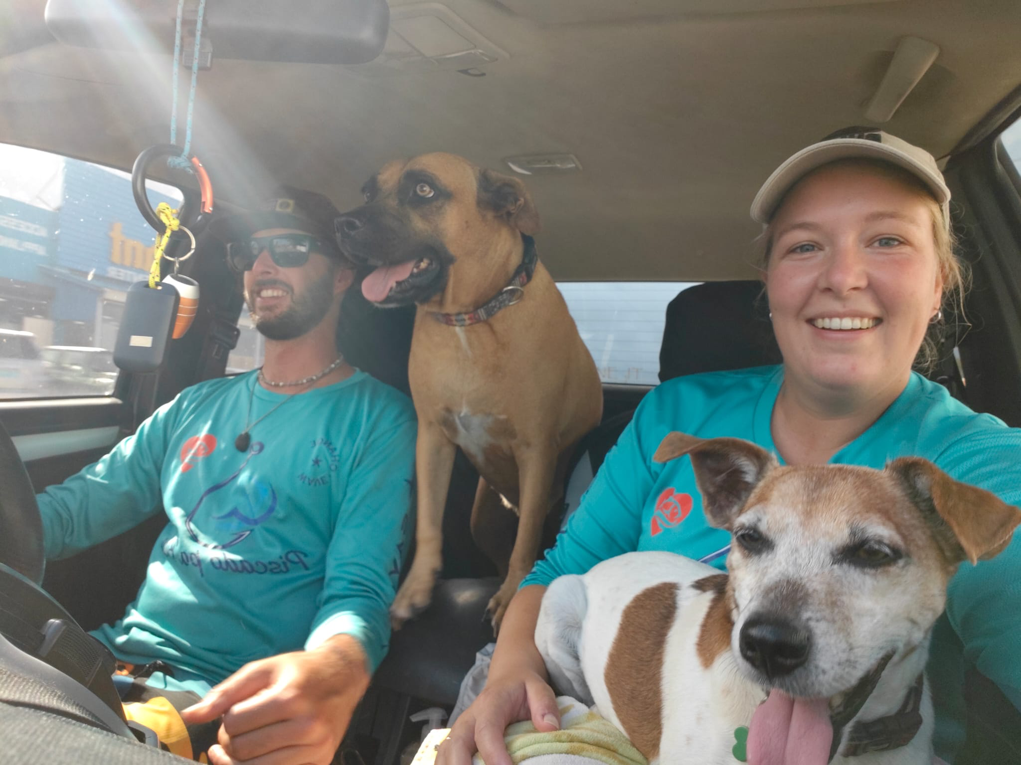 Two smiling people with dogs inside a vehicle, both wearing teal shirts. The person on the left, wearing sunglasses, has a dog with light brown fur and blue eyes sitting next to them, while the person on the right, wearing a beige cap, is holding a white and brown dog.