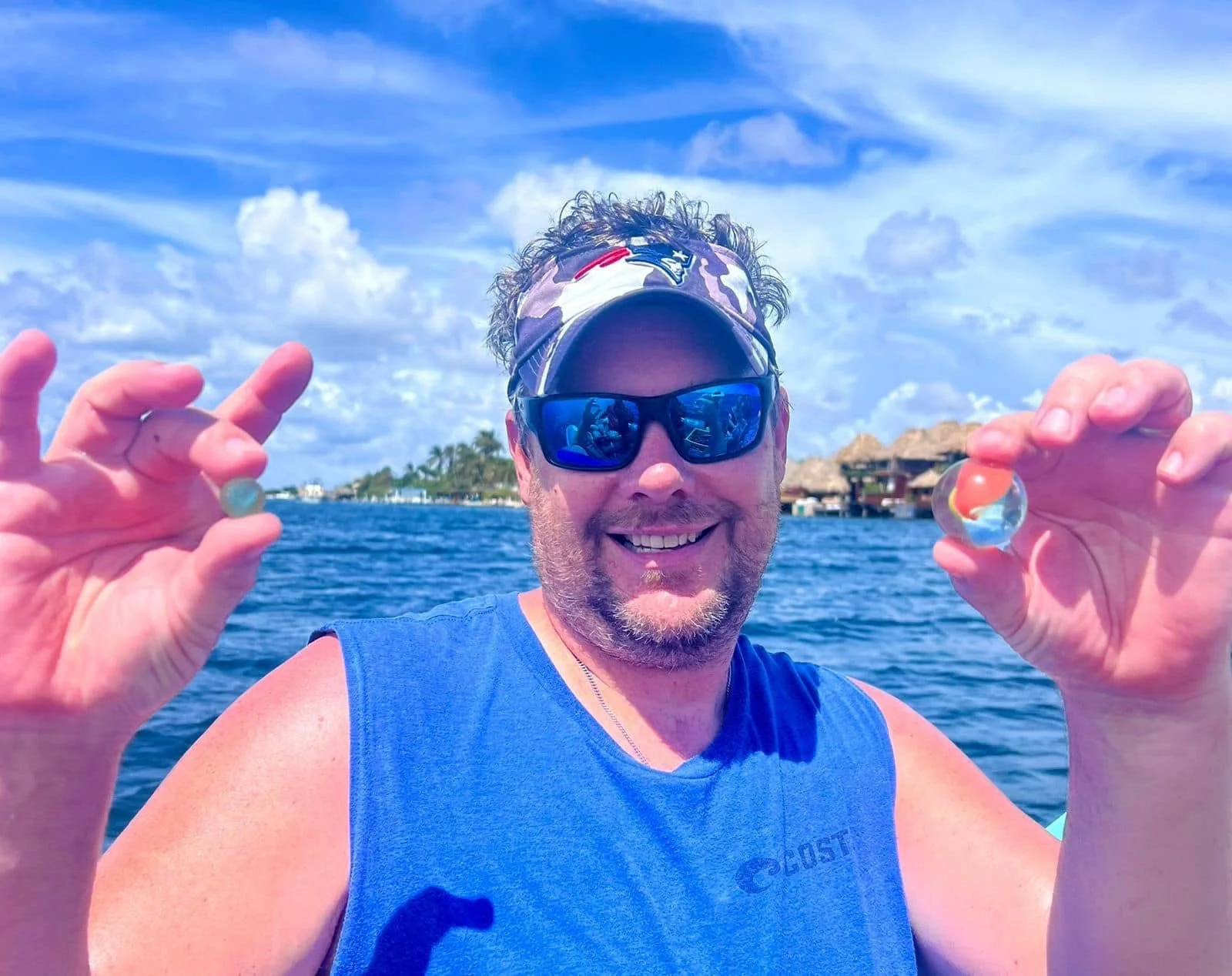 Man smiling on water with tropical scenery, holding two glass marbles.