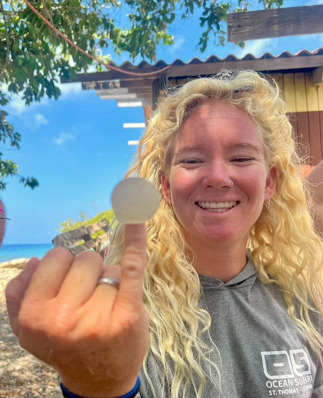 A blonde woman with curly hair smiling and showing her middle finger with a ring, standing outdoors near a beach with trees and a wooden structure in the background.