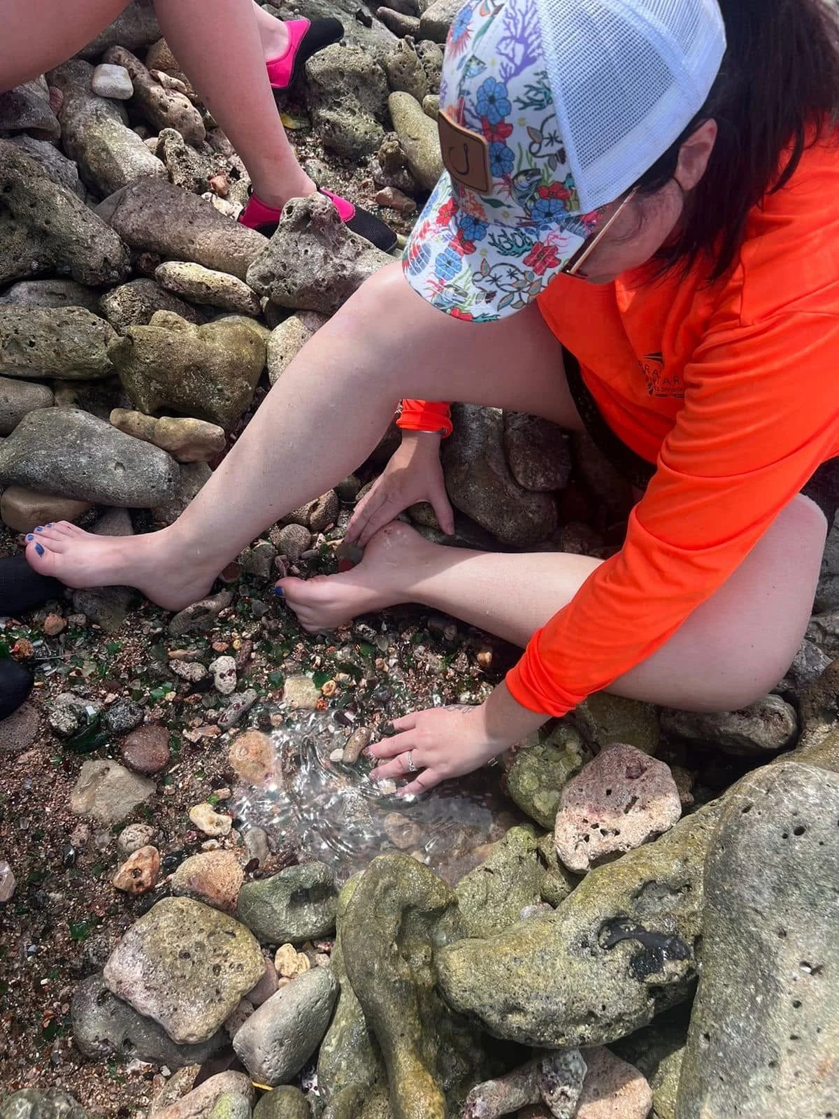 A person with a floral cap and orange jacket is sitting on a rocky shore, reaching into a small tide pool surrounded by large and small rocks, with another person's leg visible in the background.