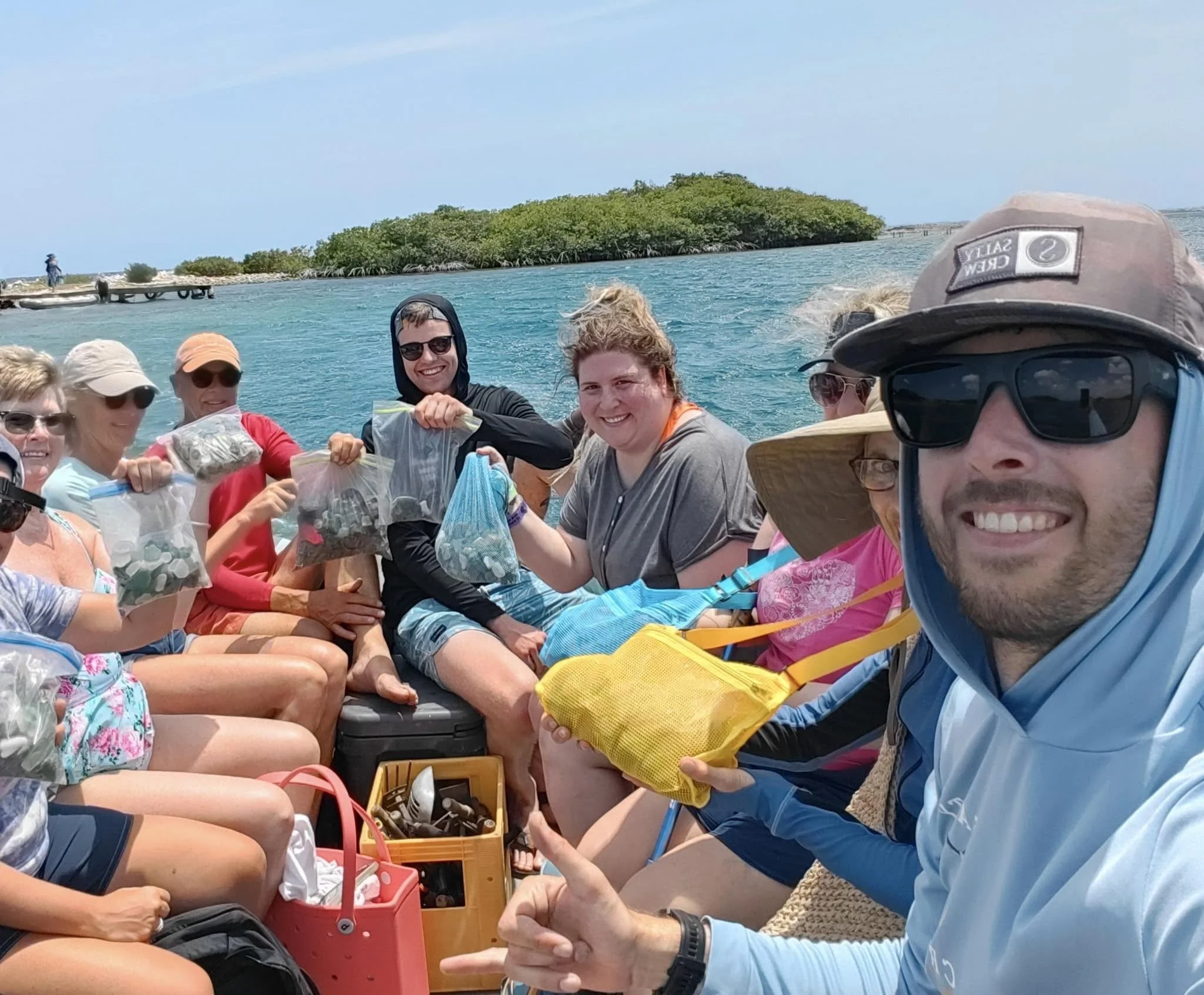 Group of people on a boat, holding bags of seashells, with some smiling at the camera, near a shoreline with green trees and a clear blue sky.