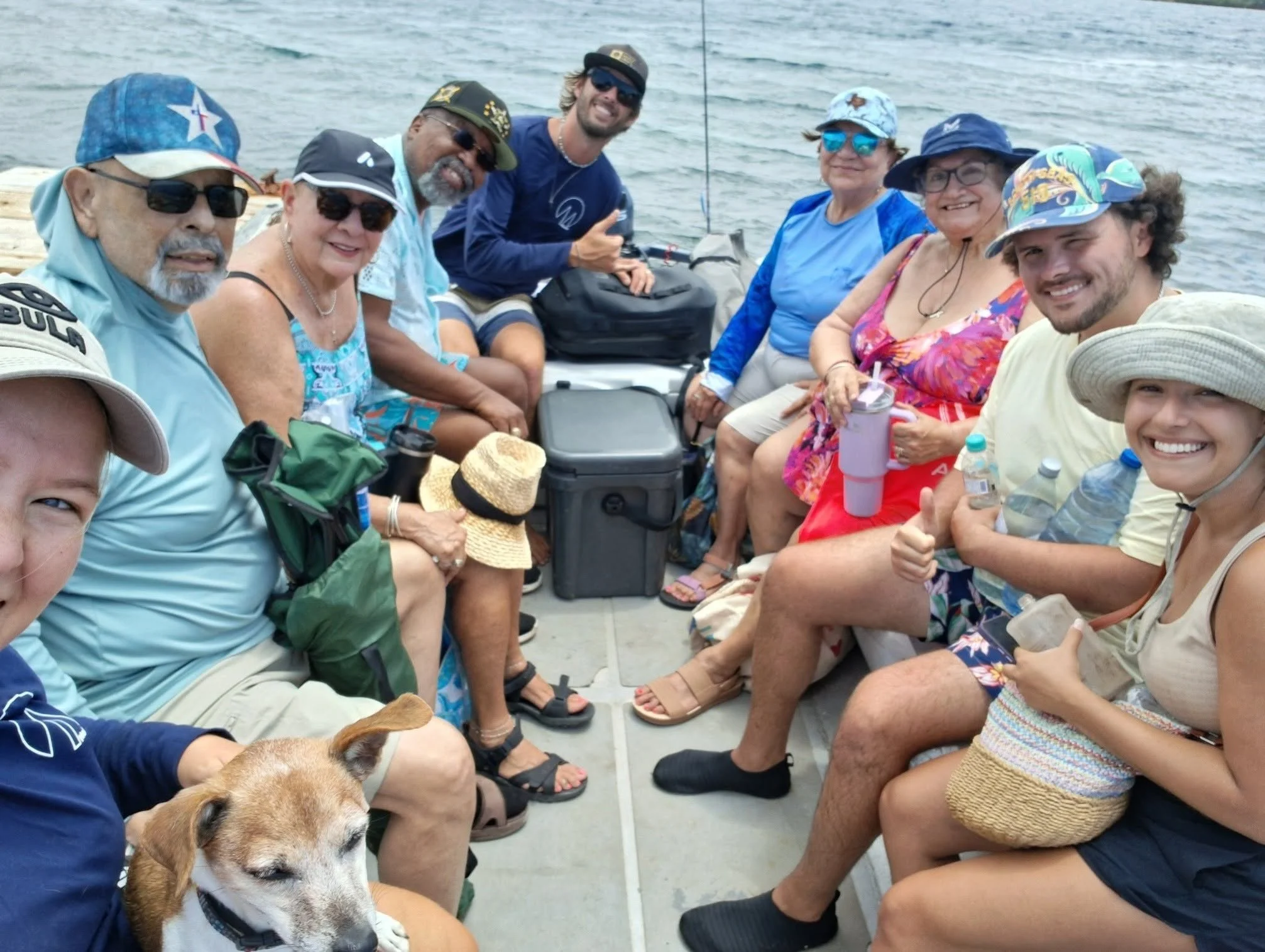 A group of people sitting on a boat deck, enjoying the day and smiling for the camera. They are wearing casual summer clothes, hats, and sunglasses, with some holding water bottles. The background shows water and the sky.