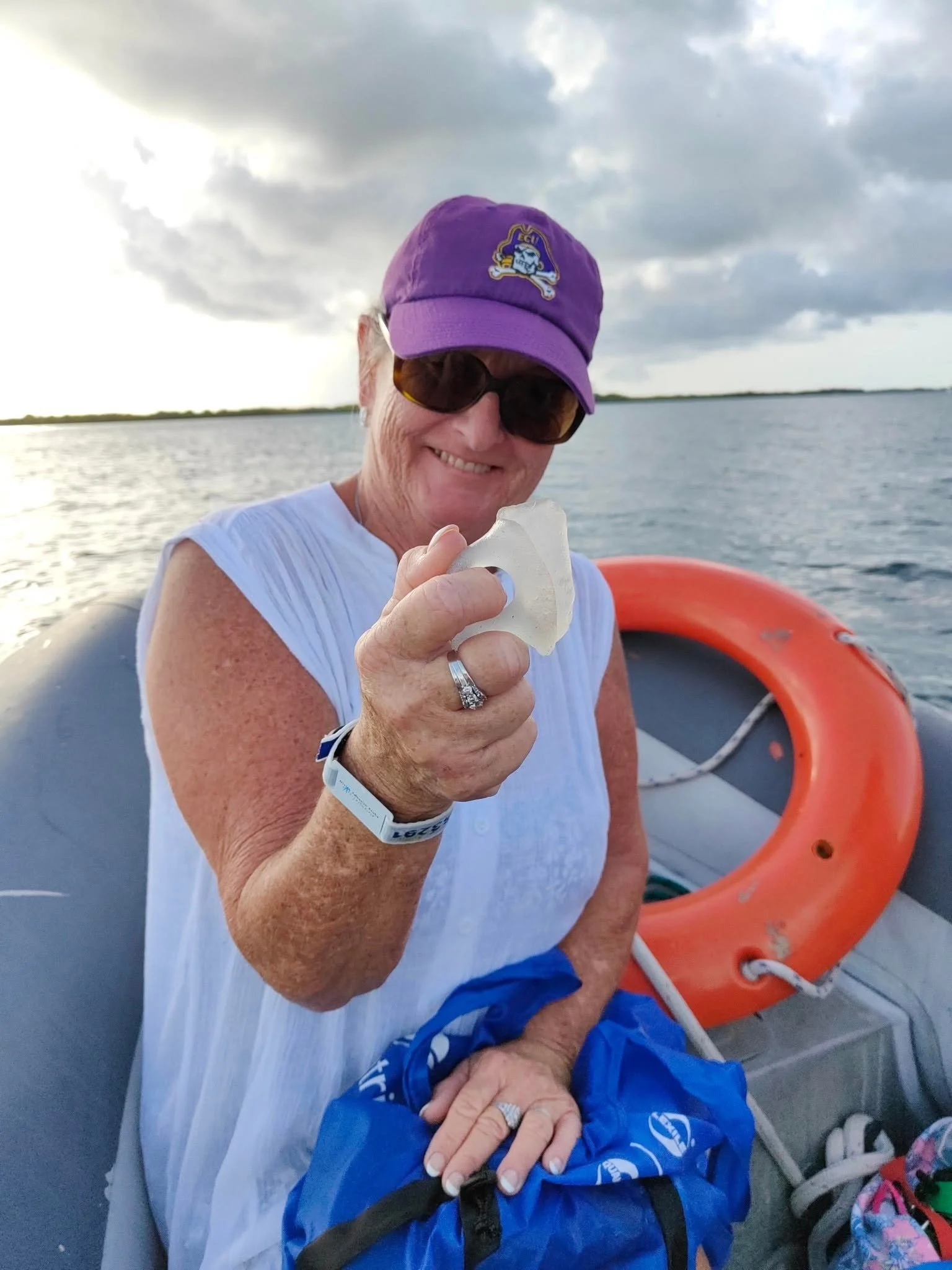 A smiling older woman wearing sunglasses and a purple cap that has a logo, holding a seashell, seated on a boat with life rings, water, and cloudy sky in the background.
