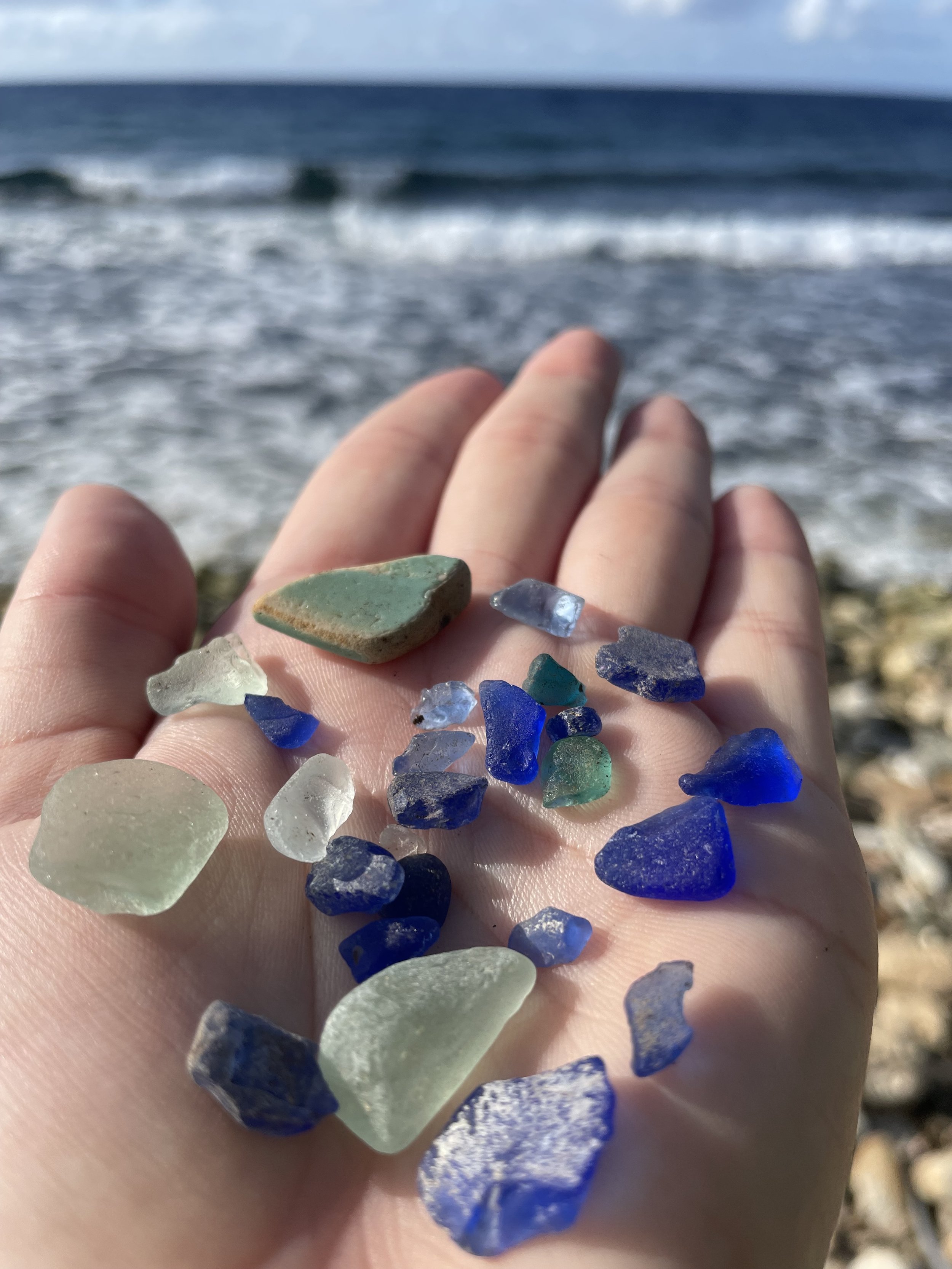 Person holding various pieces of colorful sea glass and small shells on a beach with ocean waves in the background.
