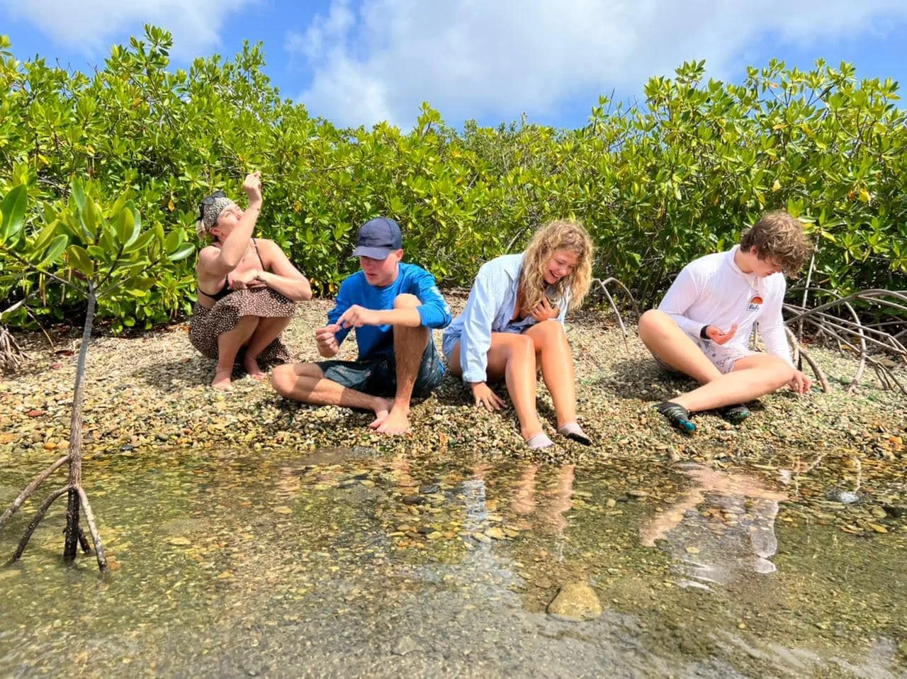 Four children sitting on a shoreline filled with sea glass near green bushes, laughing and playing.