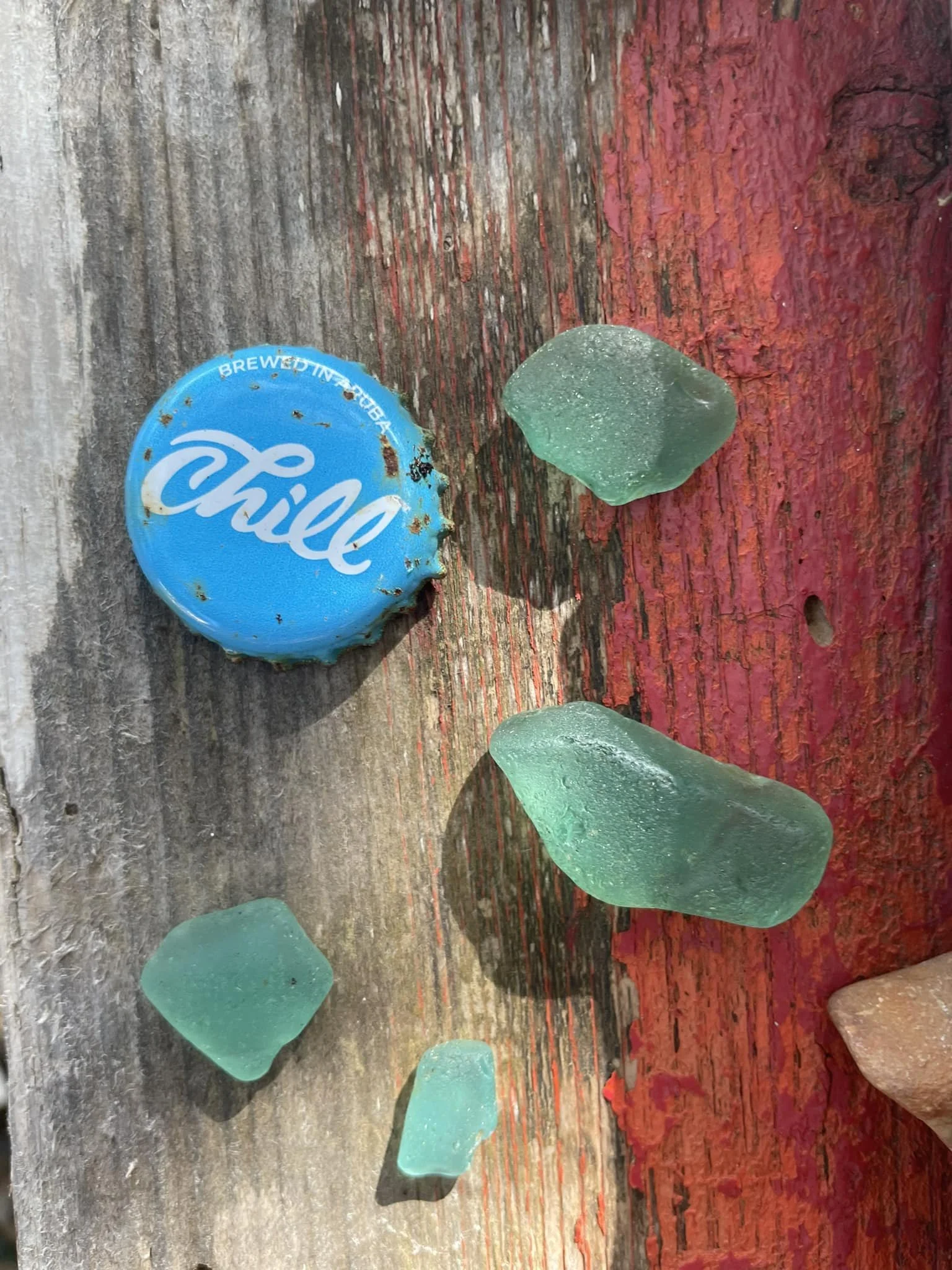 Piece of teal sea glass, old blue bottle cap with white lettering and rust spots, and some red and green colored sea glass on a weathered wooden surface with reddish paint.