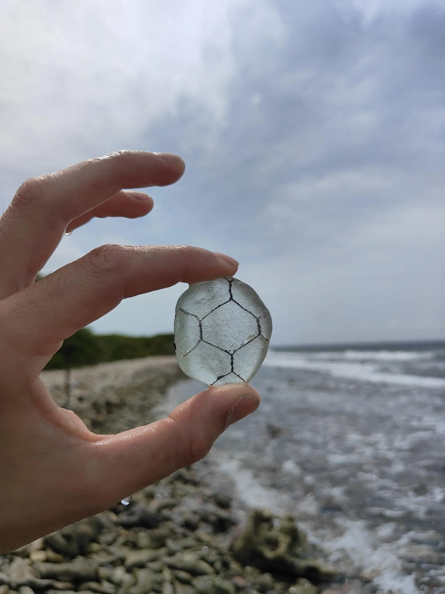 A hand holding a shattered piece of white glass with black crack lines, with a cloudy sky and a rocky shoreline and water in the background.