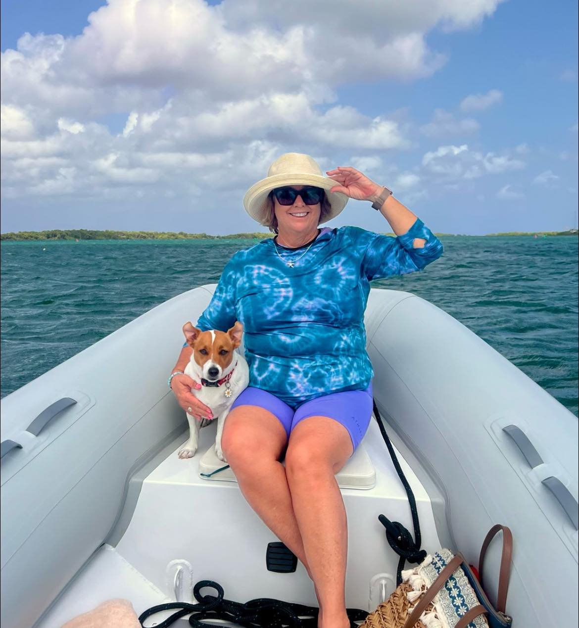 Woman in a blue tie-dye shirt and purple shorts, wearing a sun hat and sunglasses, sitting on a boat with a small dog, on a body of water with land in the background.
