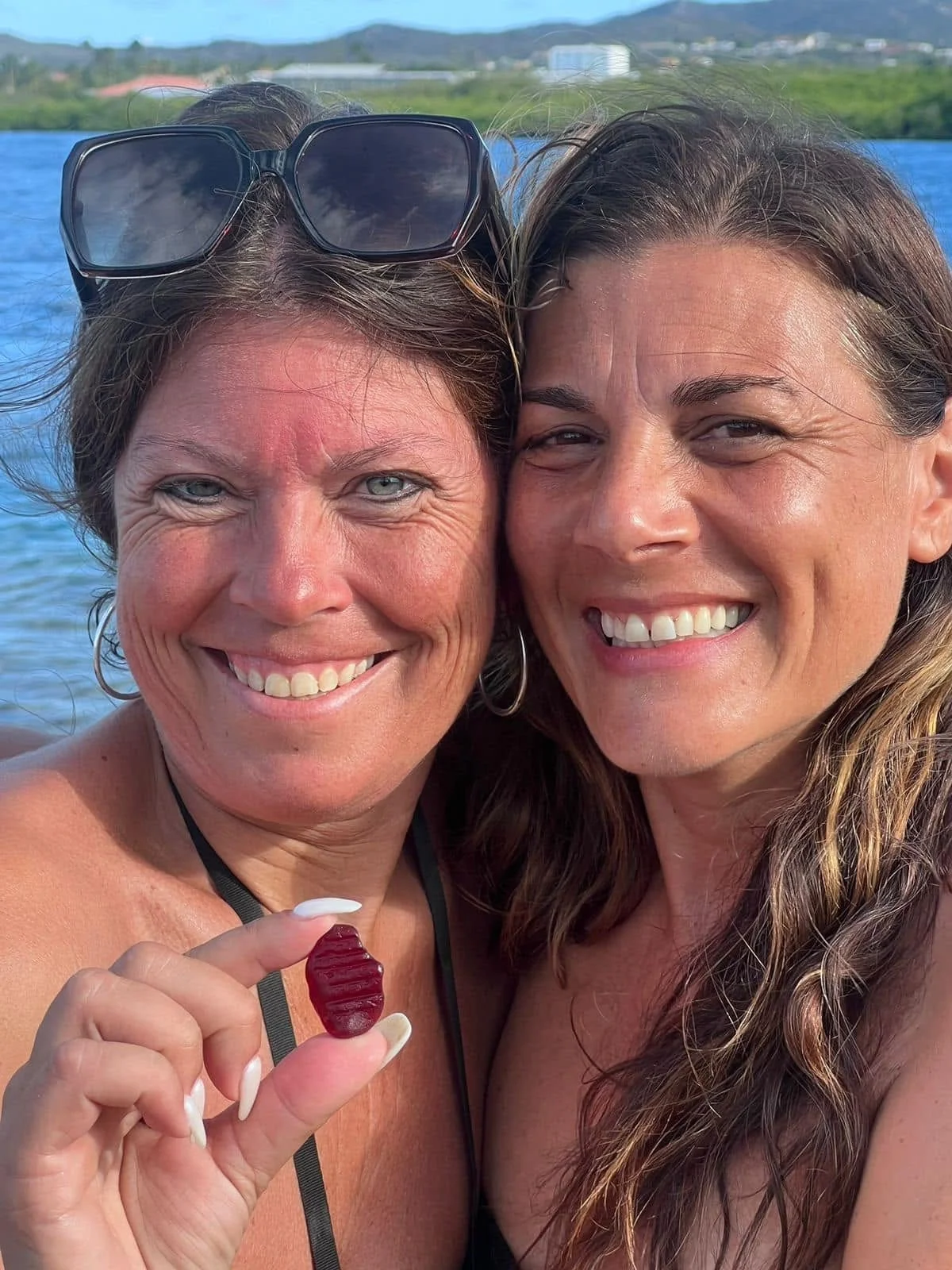Two women close together smiling by a body of water, with a landscape and distant buildings in the background. One woman is holding a small, red gummy candy.