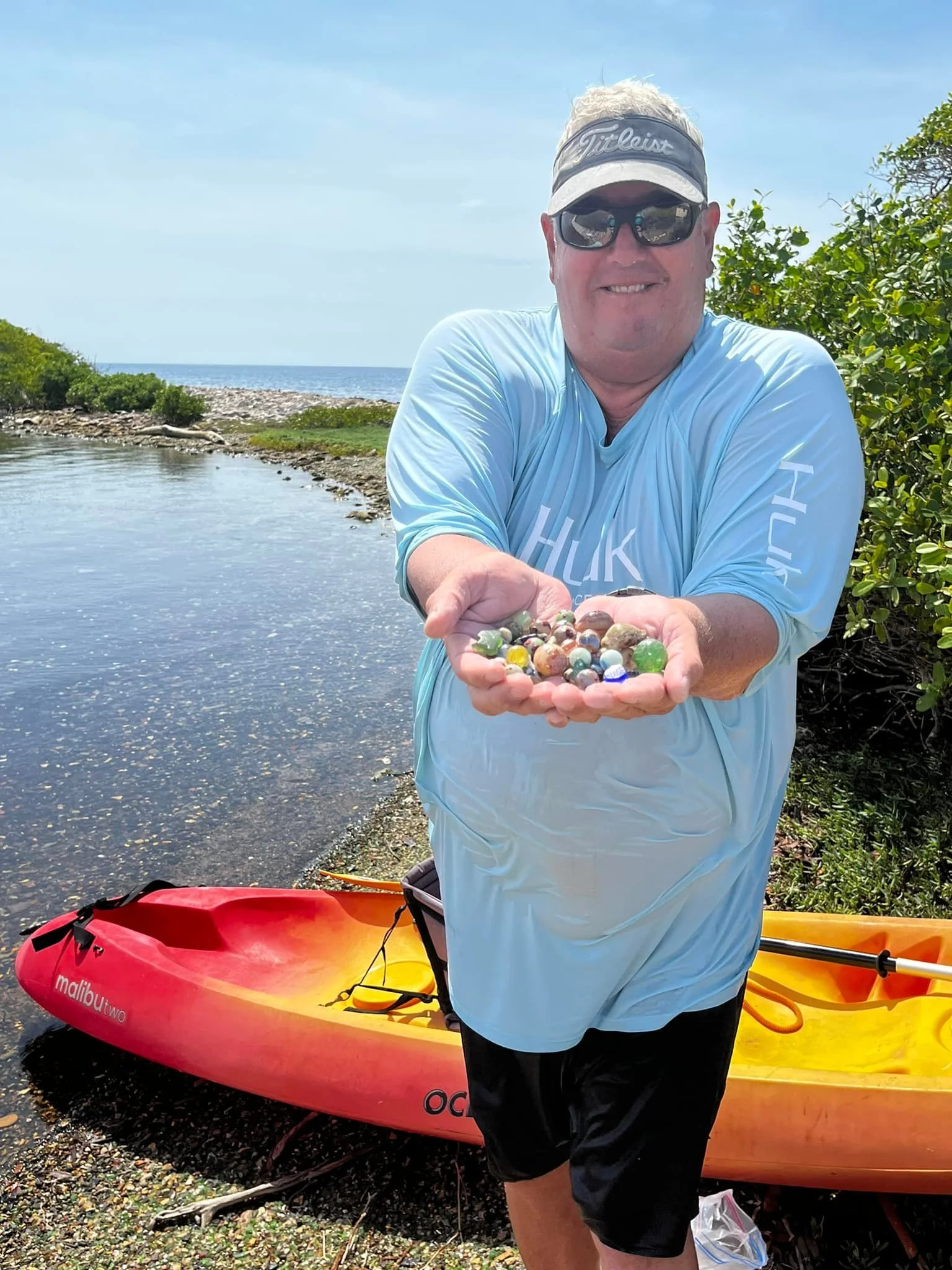 Man standing outdoors near water with two kayaks, holding and presenting colorful sea glass.