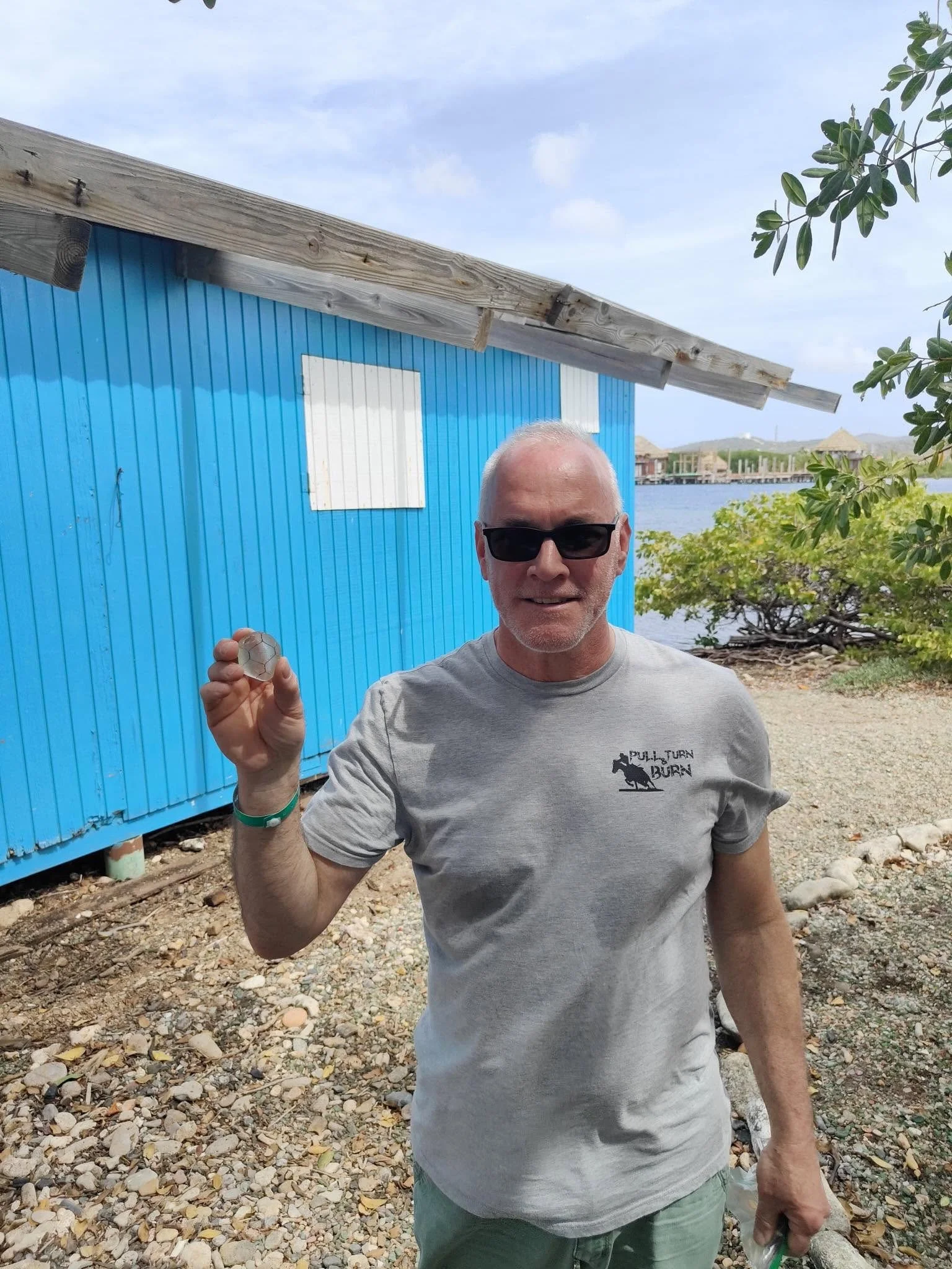 A smiling man wearing sunglasses and a gray t-shirt with a logo, holding a small object in his right hand, standing outdoors on a pebbled ground near a blue wooden building with a slanted roof, with trees and water in the background.