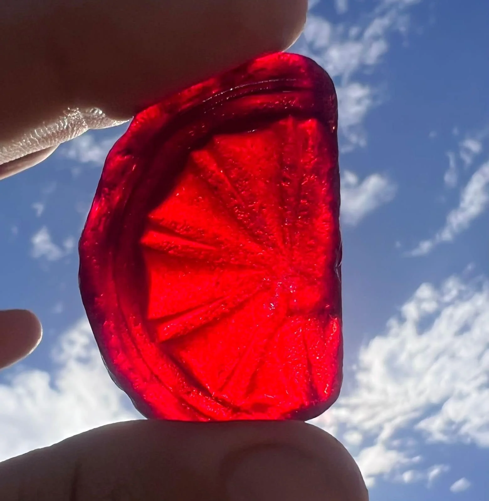 Close-up of a red piece of sea glass held between fingers against a bright sky with scattered clouds.