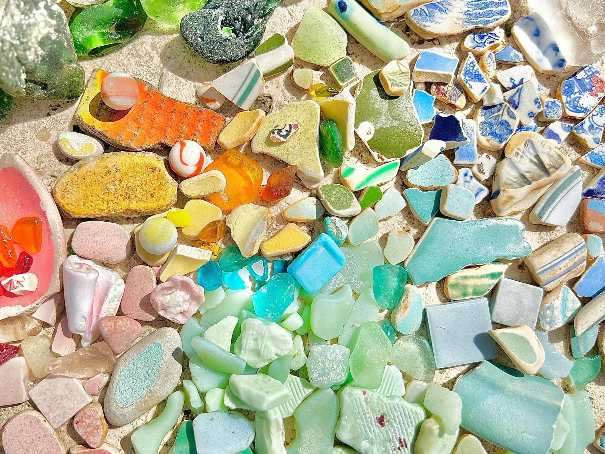 Colorful pieces of sea glass scattered on a sandy surface, with shades of green, blue, white, pink, orange, and yellow.