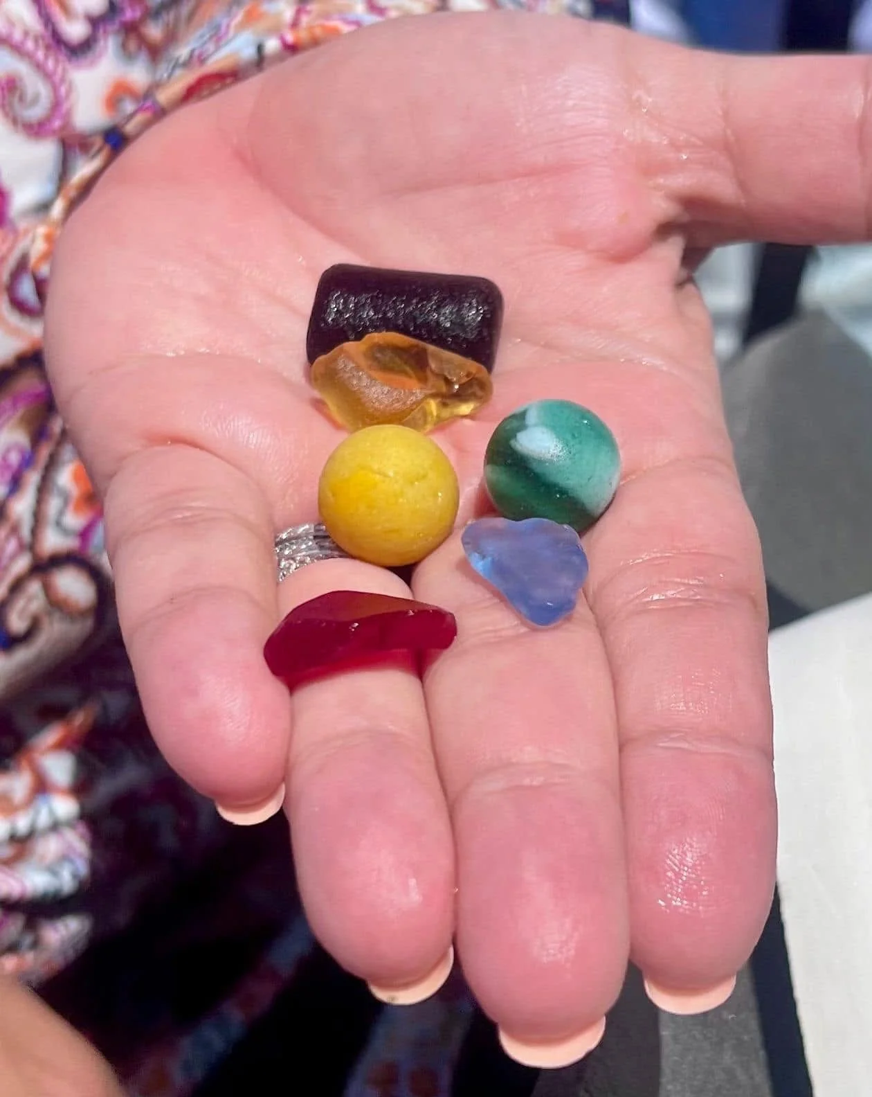 A person's hand holding seven small, colorful pieces of sea glass of various shapes and colors.
