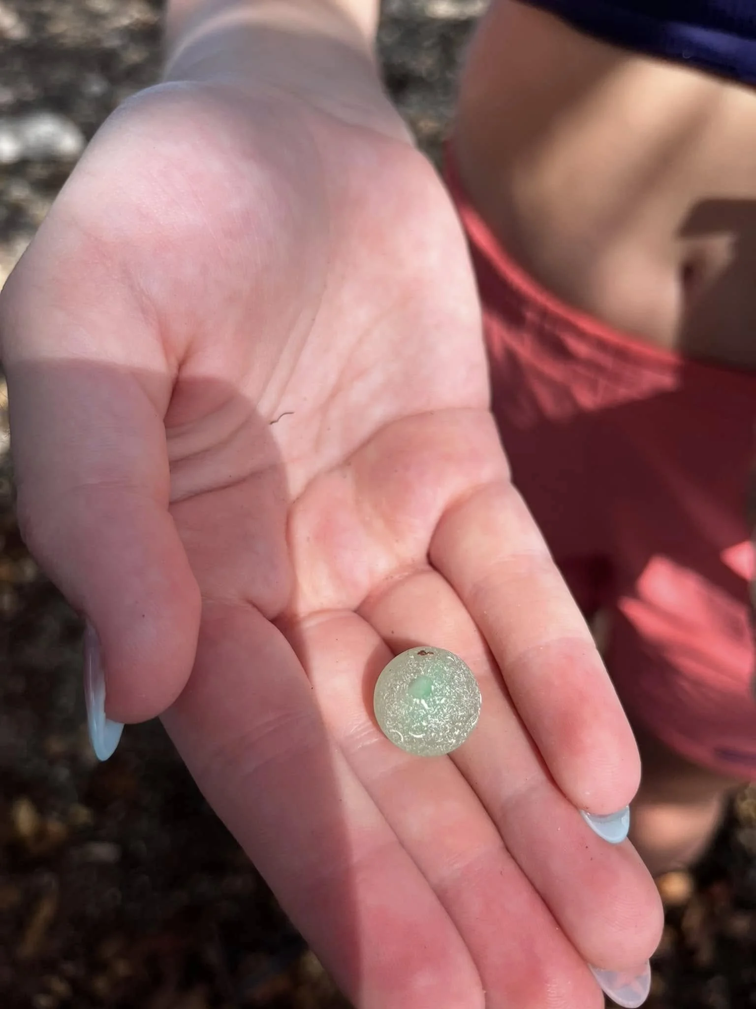 Close-up of a person's hand holding a small, round, shiny greenish globe or mineral. The background appears to be dirt or soil, and part of another person's clothing is visible.
