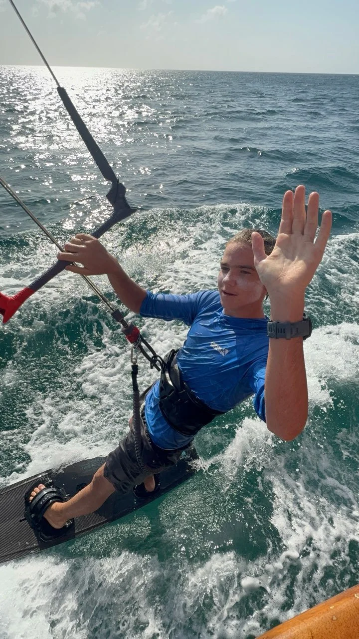 A man in a blue wetsuit and riding a wakeboard on the ocean, holding onto a kite control bar, with the sea and sky in the background, waving with his right hand.