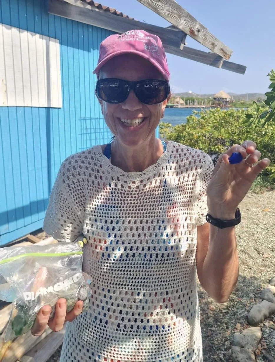 A smiling woman wearing sunglasses, a pink cap, and a white mesh top, holding a bag of gemstones and a small blue object, outdoors near water with greenery and structures in the background.