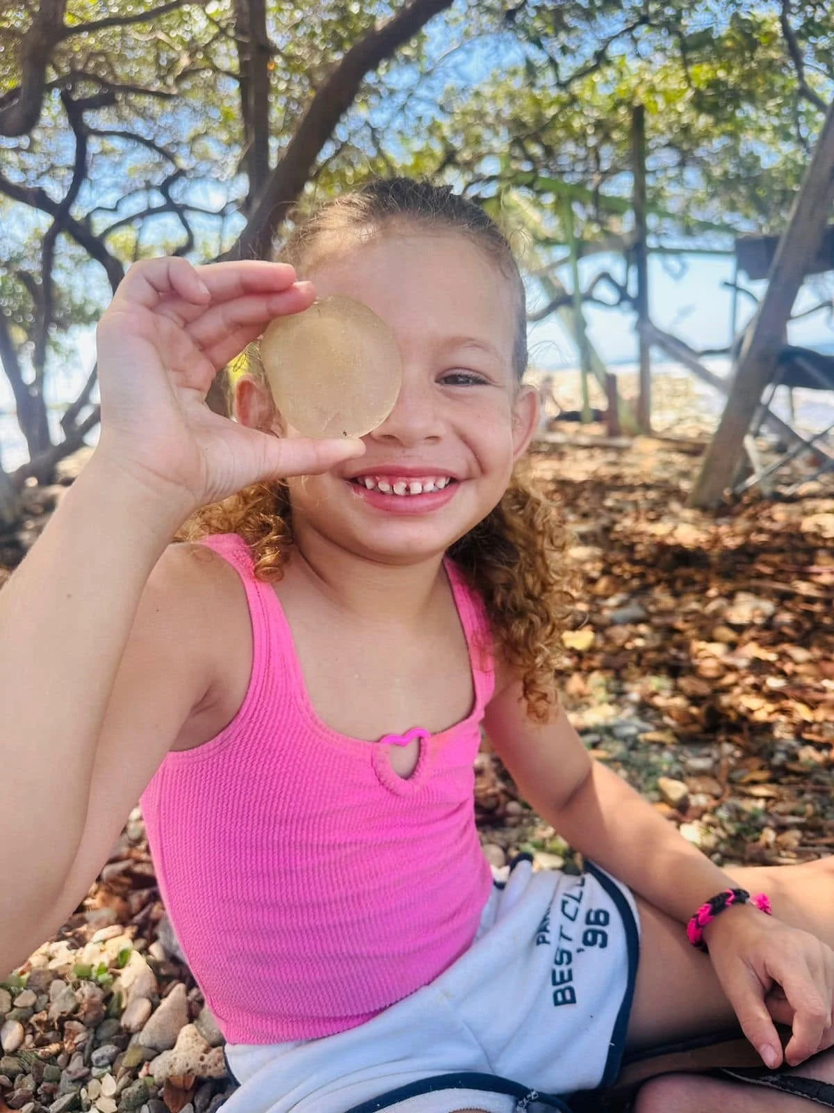 A young girl with curly hair smiling and holding a translucent, round object up to her face outdoors. She is wearing a pink tank top and white shorts, sitting on a rocky ground under a tree with sunlight filtering through the leaves.