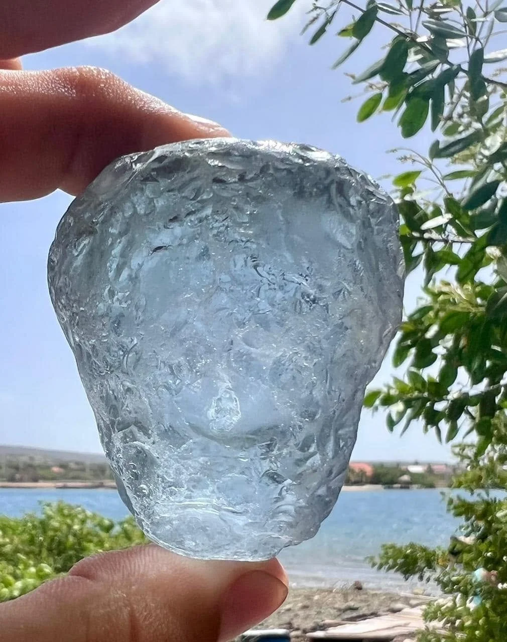 A person holds a large, irregularly shaped ice chunk outdoors with a body of water, trees, and a partly cloudy sky in the background.