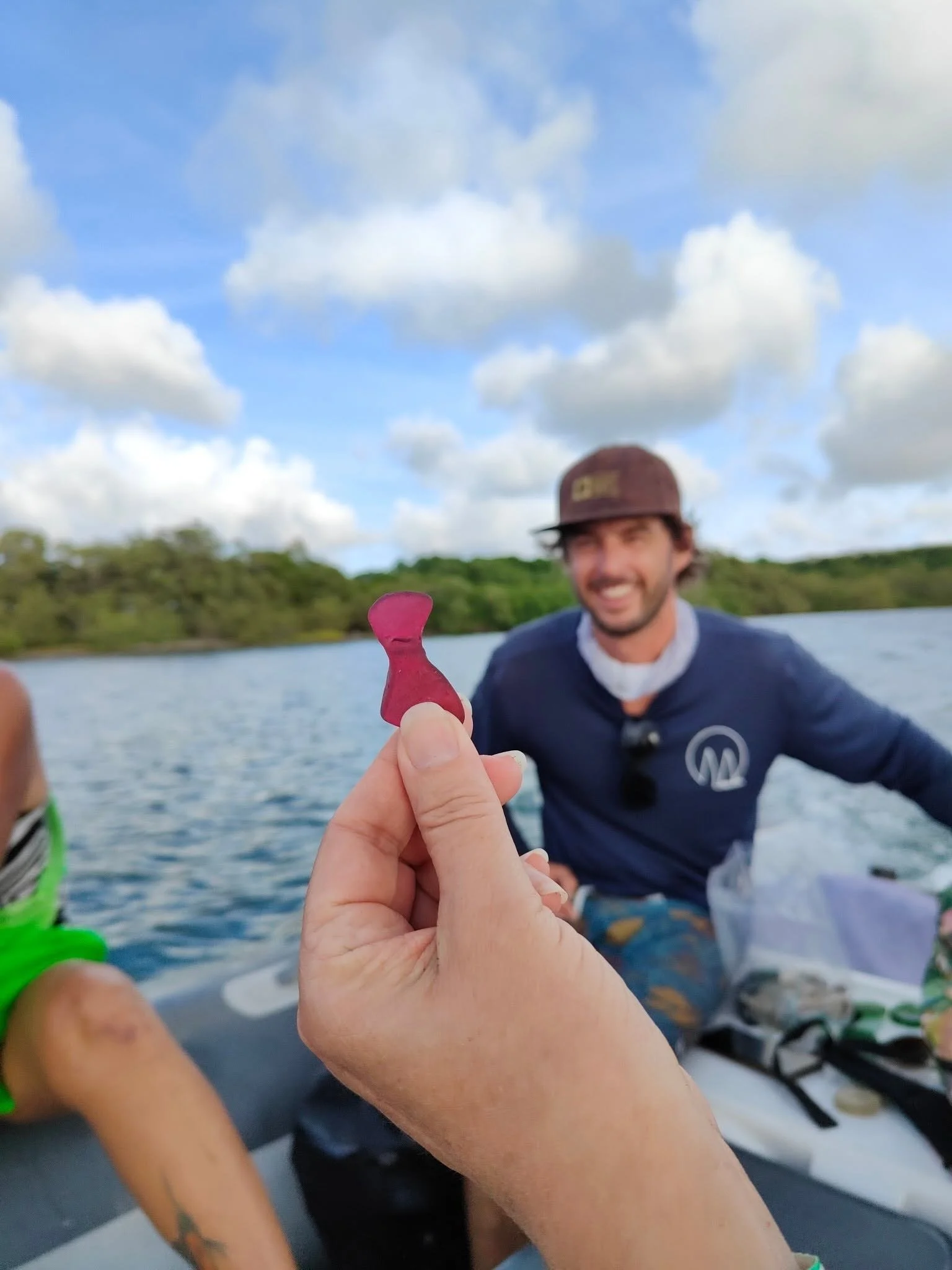 Person smiling and holding a small pink dog-shaped gummy candy in front of the camera on a boat, with water, trees, and cloudy sky in the background.