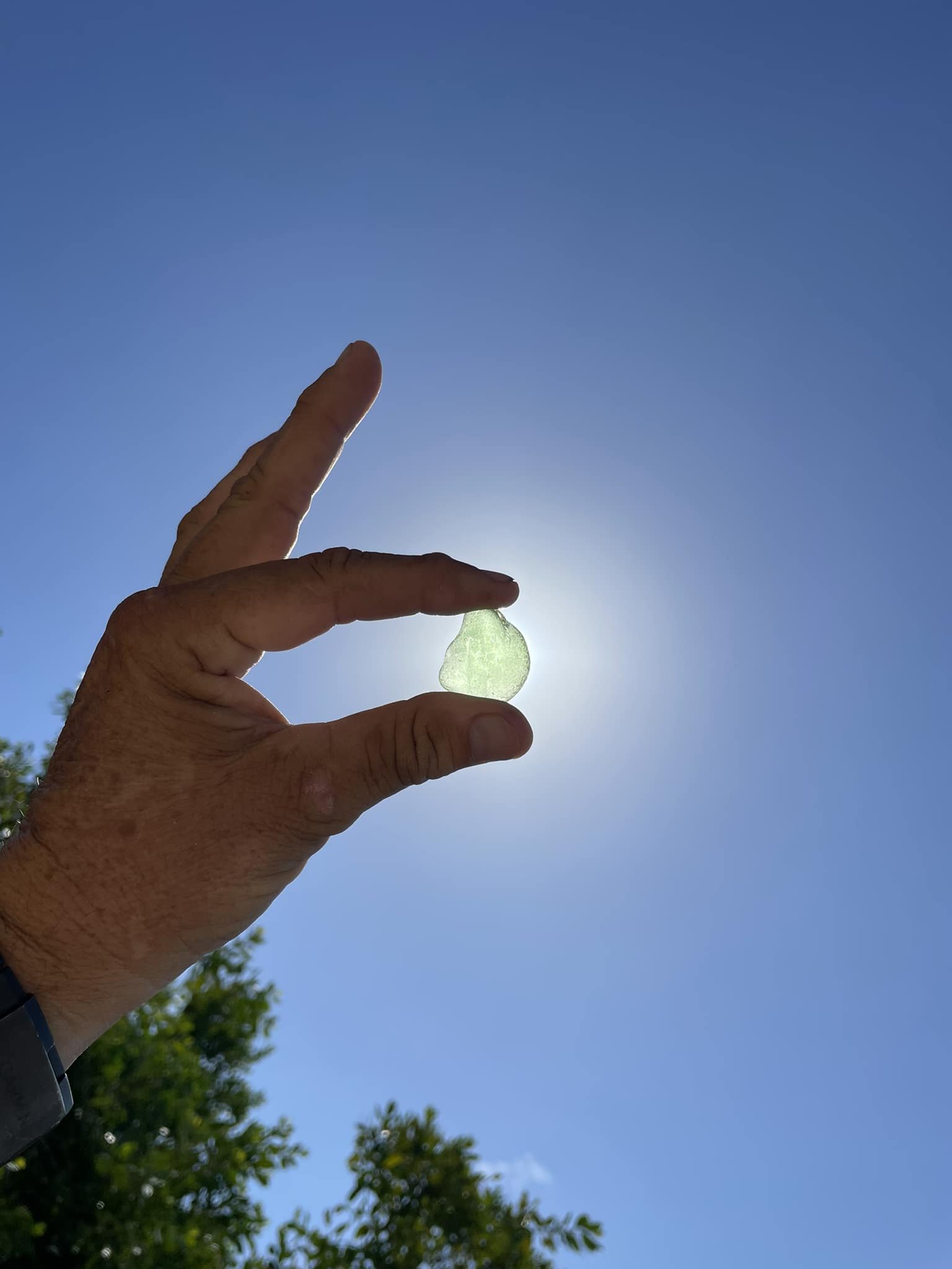 A hand holding a piece of translucent green jade against a bright blue sky with some green trees in the background.