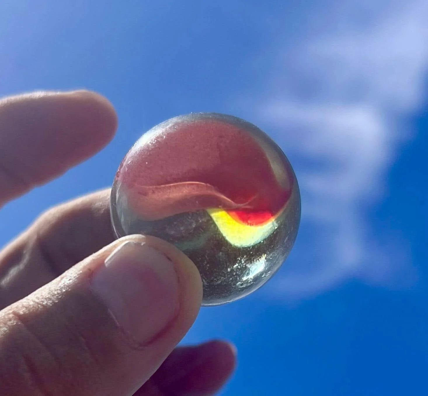 Close-up of a hand holding a small glass marble up to the sky, showing colorful reflections and a background of a blue sky with some clouds.