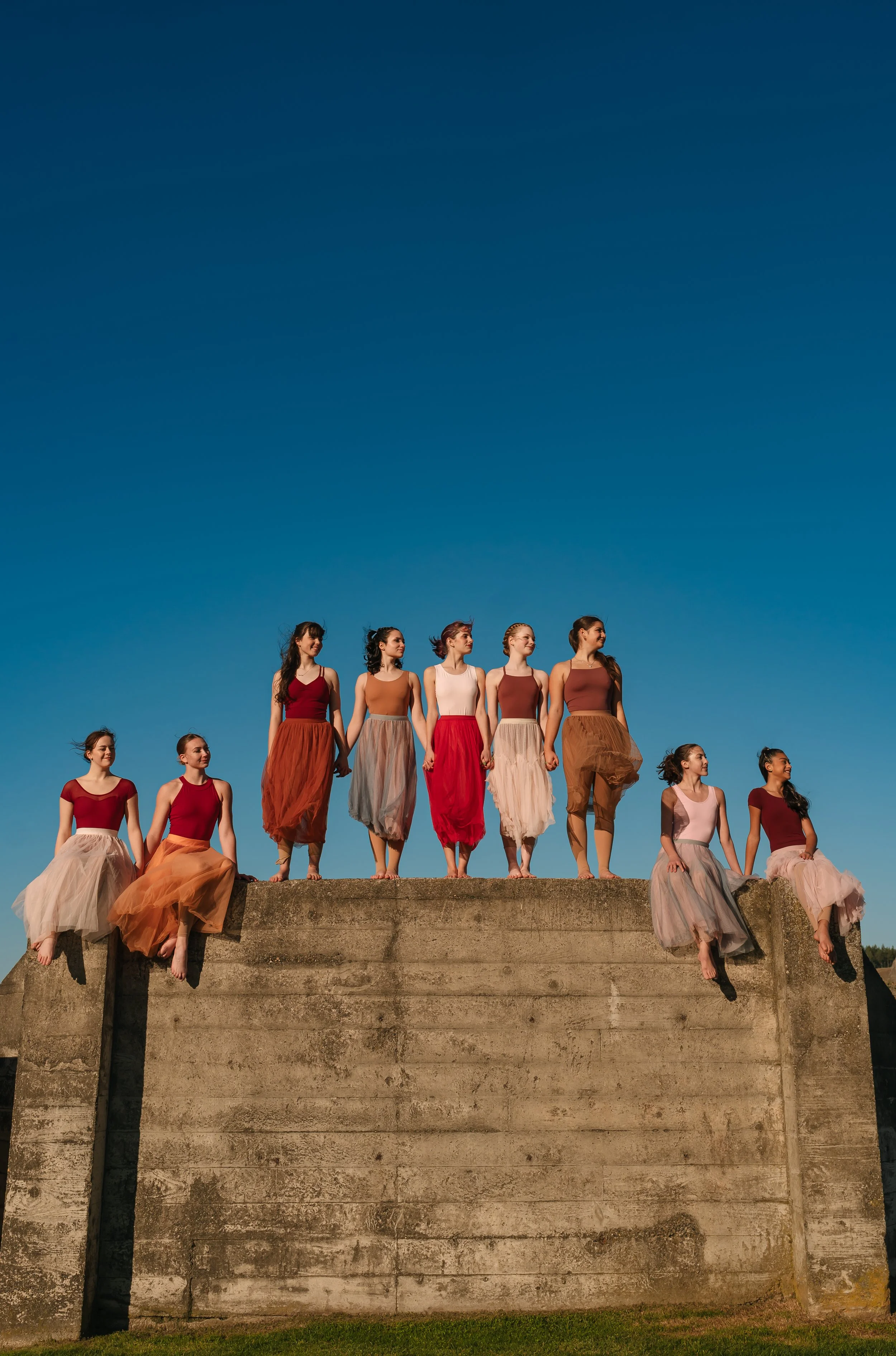 A group of dancers wearing colorful skirts and tops stand barefoot on a concrete wall against a clear blue sky.