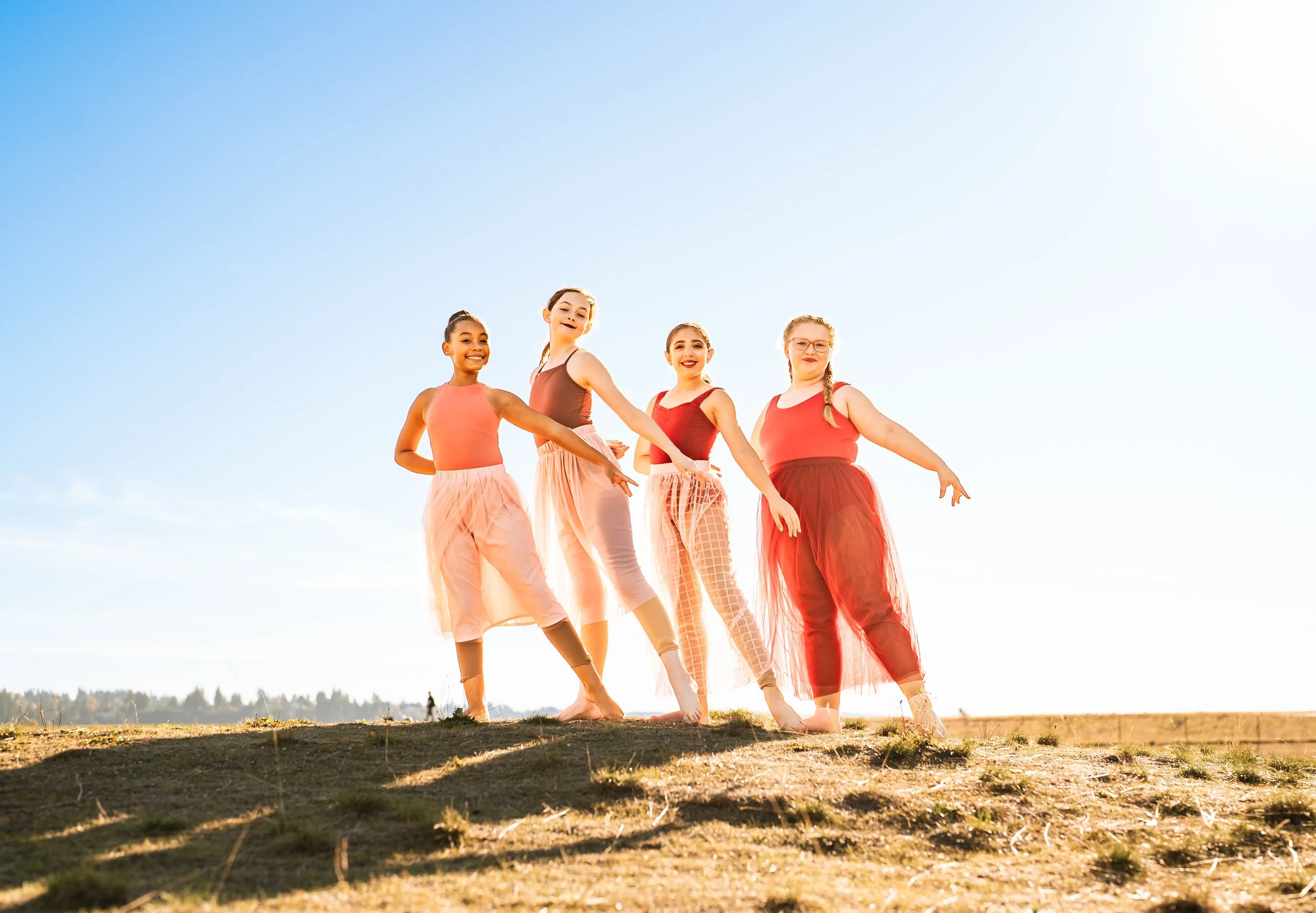 Four dancers in red and pink outfits posing outdoors on a sunny day with a clear blue sky.