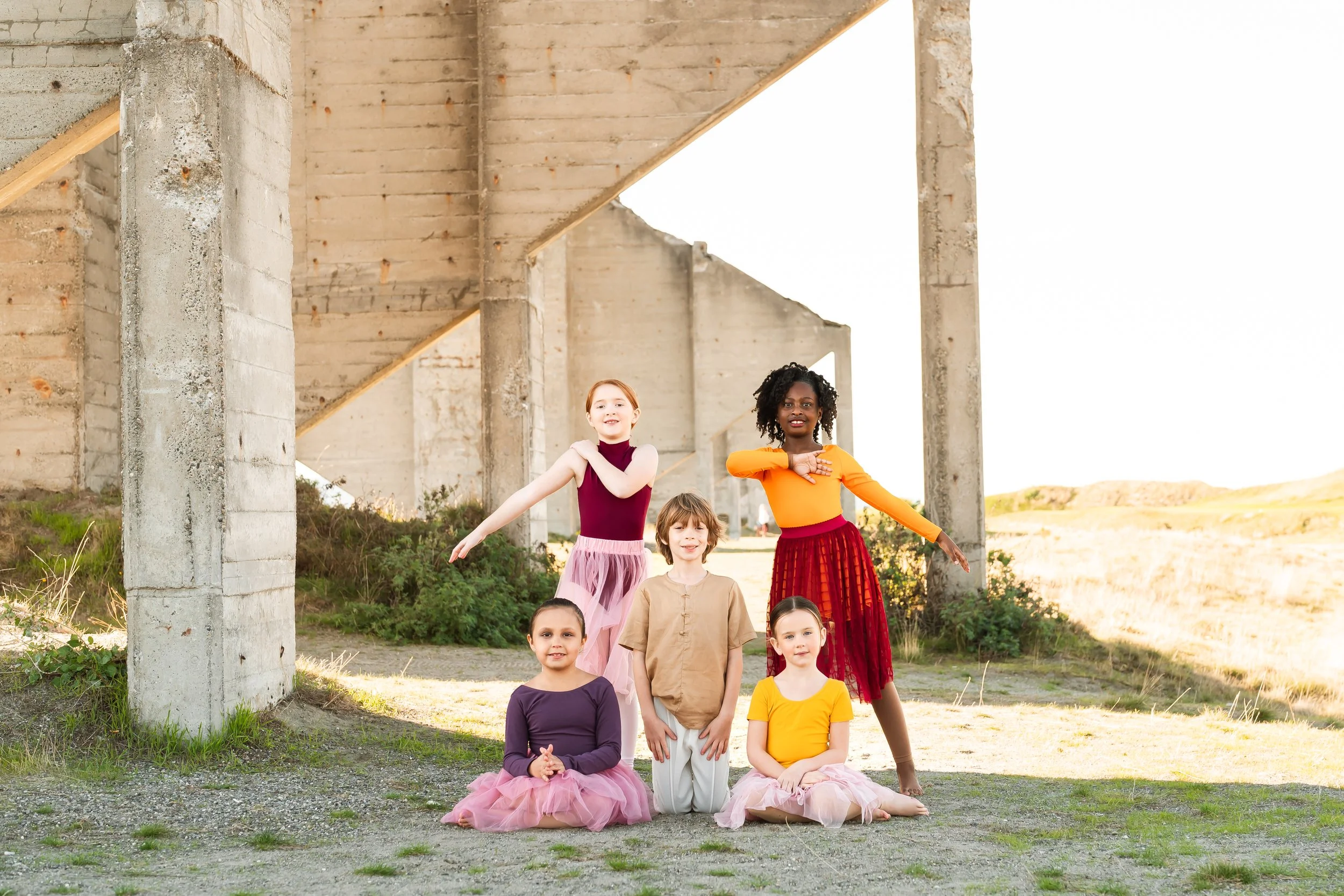 Five children in colorful dance attire posing outdoors under a concrete structure. Two children are kneeling, and three are standing, with arms gracefully extended.