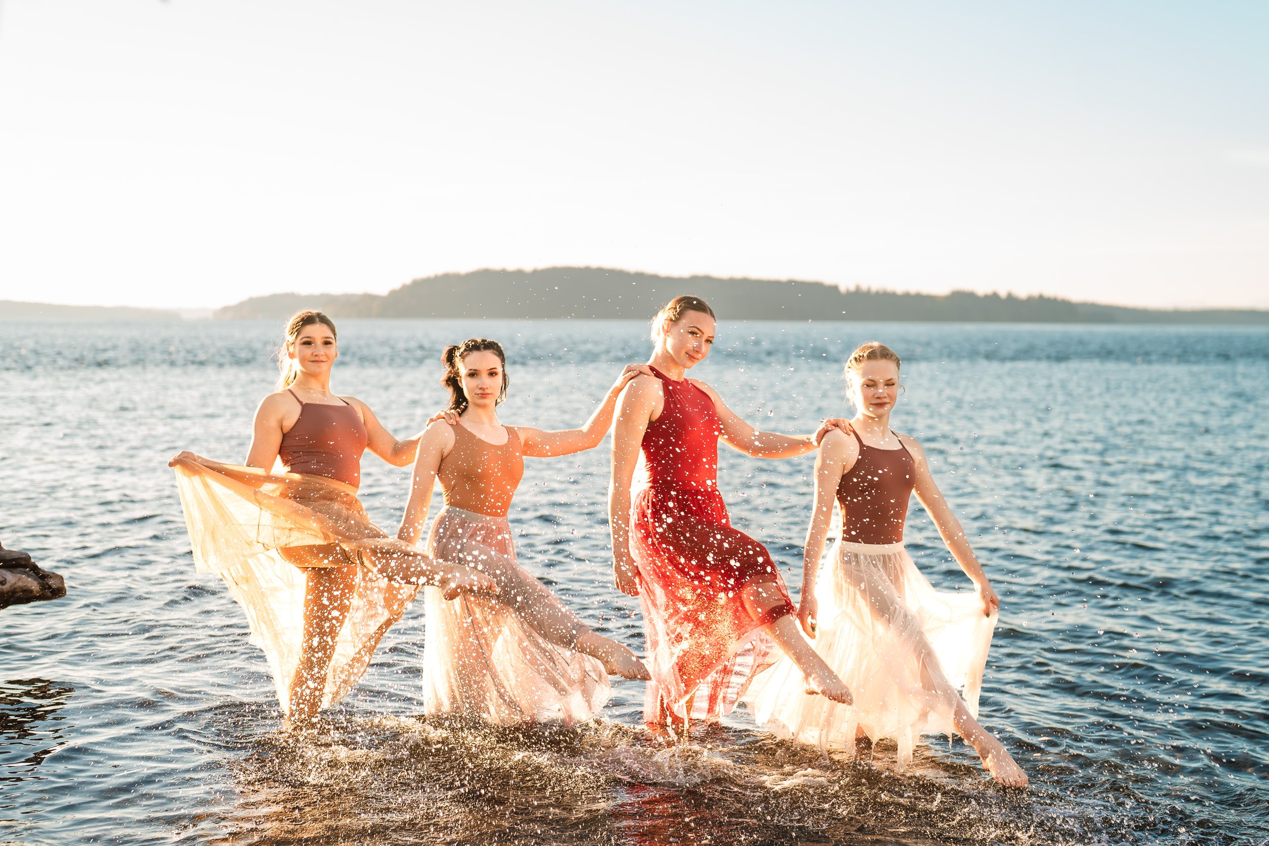 Four dancers in flowing dresses posing in shallow water with splashes, against a scenic lake background during sunset.