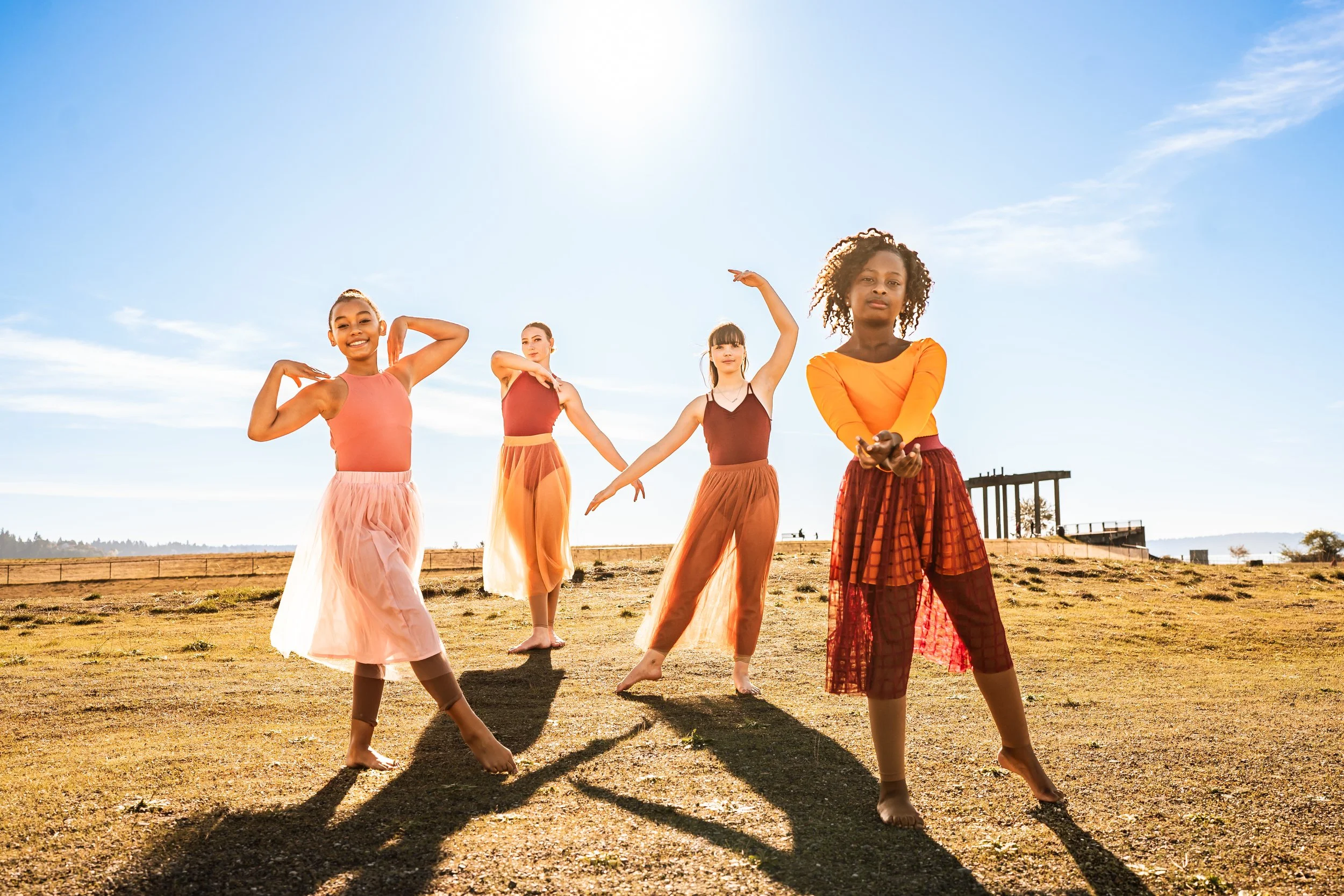 Four dancers posing outdoors under a clear blue sky, wearing colorful skirts and tops.