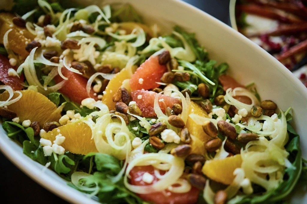Close-up of a salad featuring greens, sliced tomatoes, orange slices, shredded cheese, and sunflower seeds.