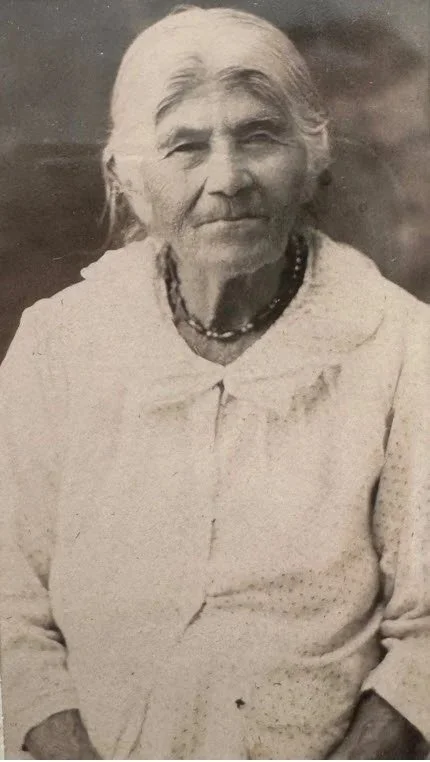 A black-and-white photo of an elderly woman with gray hair tied back, wearing a light-colored dress and a beaded necklace, standing outdoors.