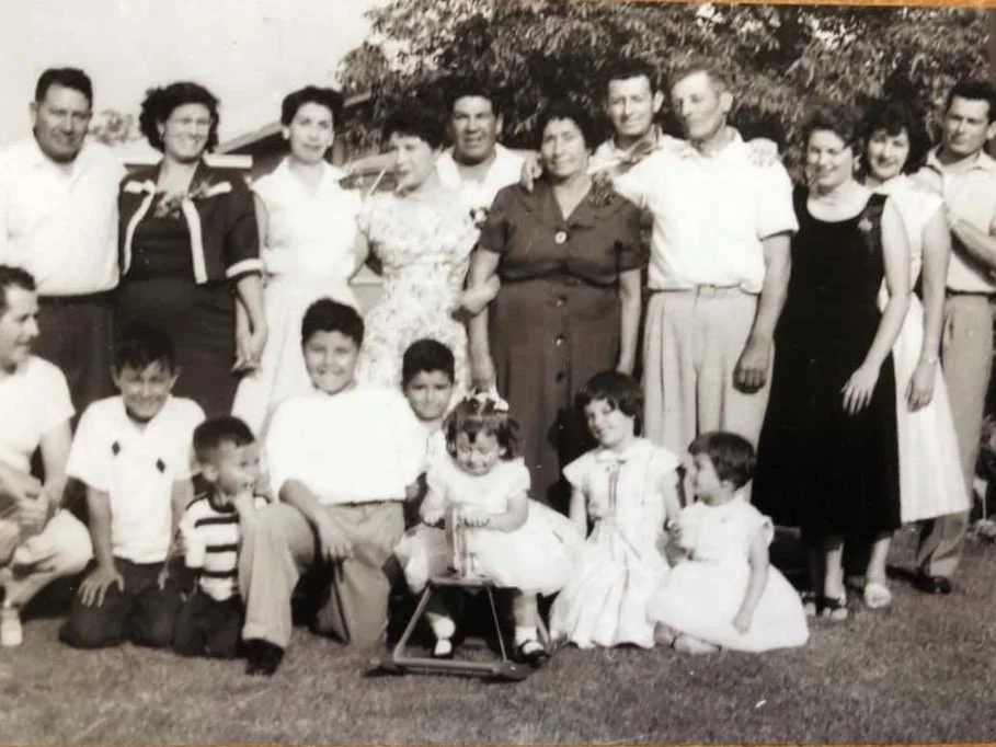 A black and white photo of a large family group of men, women, and children outdoors, standing and sitting on the grass, some smiling and looking at the camera, with trees and a house in the background.