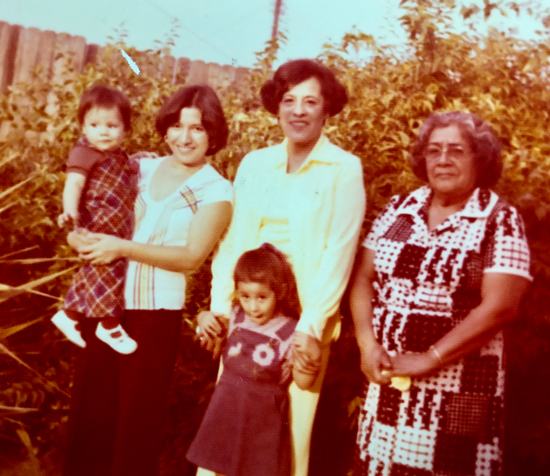A family of five women and two young girls standing outdoors in front of a wooden fence and yellow foliage.
