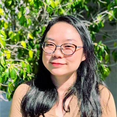 Juhyun Šakota is a Six Leaf Design client and co-principal for a project for People's Health Solutions. She is a young woman with glasses and long black hair outdoors with green foliage in the background.