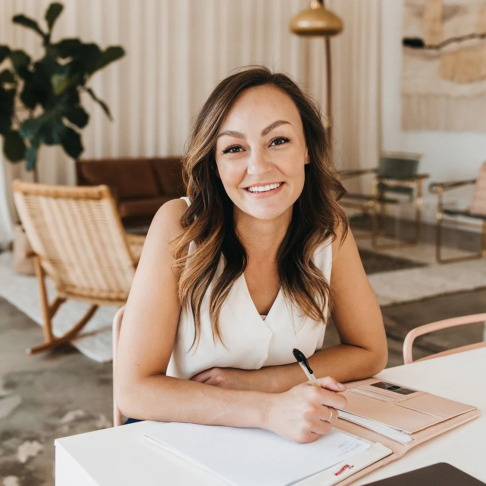 Taylor Williams is a Six Leaf Design client in Denver and a young woman with wavy brown hair and a white sleeveless top, smiling at the camera while sitting at a white table with a notebook and pen in her hand.